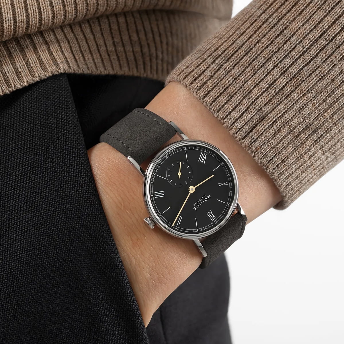 NOMOS Glashütte © 2026 Eine Person trägt eine schwarze Uhr, die an die NOMOS Ludwig erinnert, mit schwarzem Lederarmband und römischen Ziffern, kombiniert mit einem beigen Rippenpullover und einer dunklen Hose. Auf dem Zifferblatt steht "BODERRY".