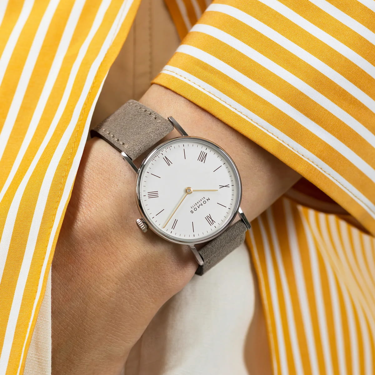 NOMOS Glashütte © 2026 Eine Person trägt eine NOMOS-Ludwig-Uhr mit weißem Zifferblatt, römischen Ziffern und einem taupefarbenen Wildlederarmband. Die Person ist mit einem gelb-weiß gestreiften Hemd bekleidet.