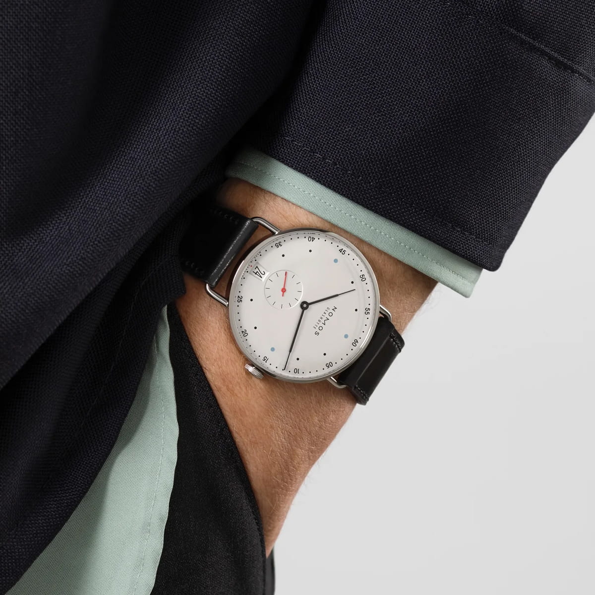 NOMOS Glashütte © 2026 Eine Person mit schwarzer Jacke und hellgrüner Hemdmanschette, die eine minimalistische NOMOS Metro-Armbanduhr mit weißem Zifferblatt, schwarzem Lederarmband und kleinem roten Zeiger trägt.