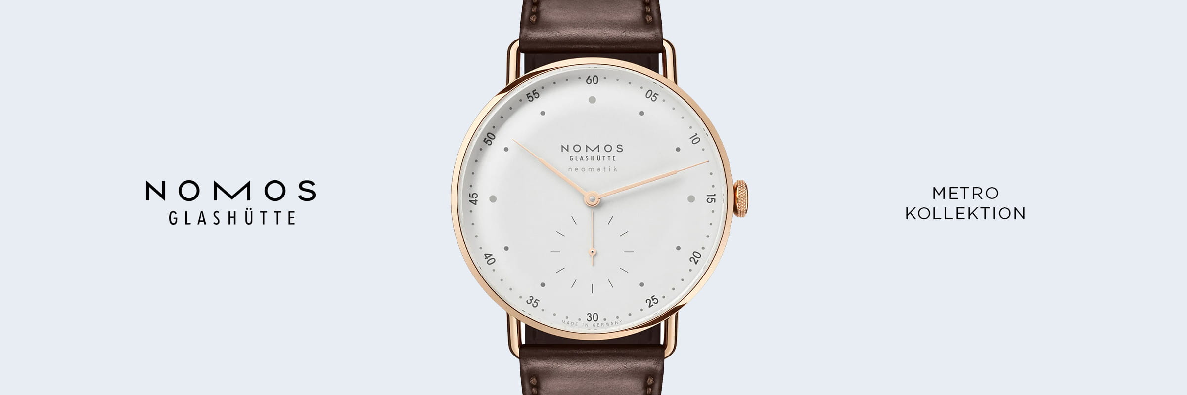 NOMOS Glashütte © 2026 Die NOMOS Metro Glashütte mit weißem Zifferblatt, goldenem Gehäuse und braunem Lederarmband. Das minimalistische Design wird in der Mitte präsentiert, seitlich stehen die Schriftzüge "NOMOS GLASHÜTTE" und "METRO KOLLEKTION".