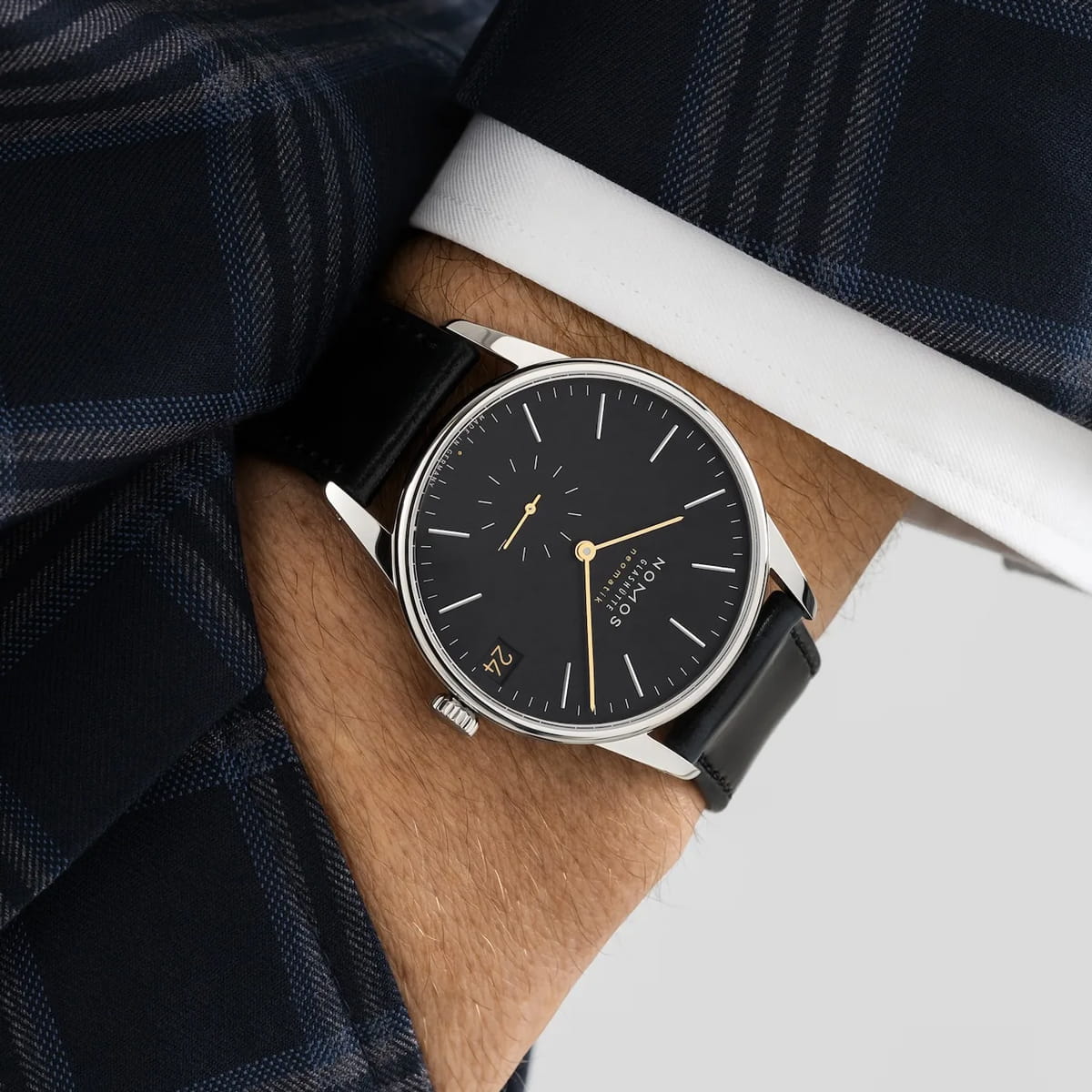 Nahaufnahme des Handgelenks einer Person, die eine schwarz-silberne Analoguhr NOMOS Orion mit schwarzem Lederarmband, minimalistischem schwarzem Zifferblatt und Datumsfenster trägt, gepaart mit einem dunkelblau karierten Anzug.