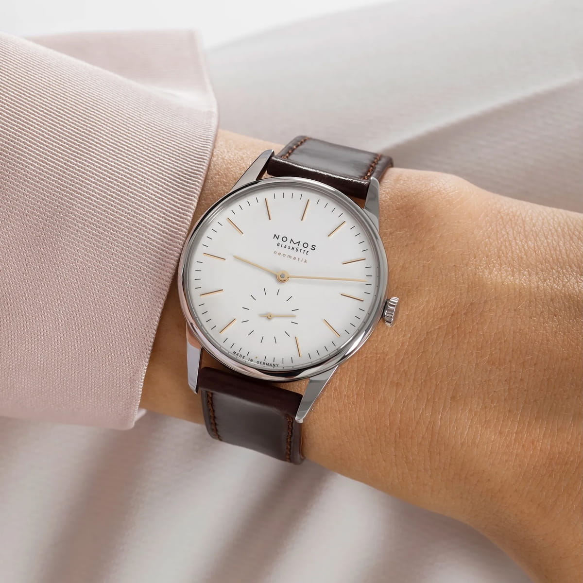 Nahaufnahme einer Person, die eine NOMOS Orion-Armbanduhr mit weißem Zifferblatt, silbernem Gehäuse, braunem Lederband und minimalistischem Design trägt. Die Person ist mit einem hellrosa Hemd mit Manschetten bekleidet.