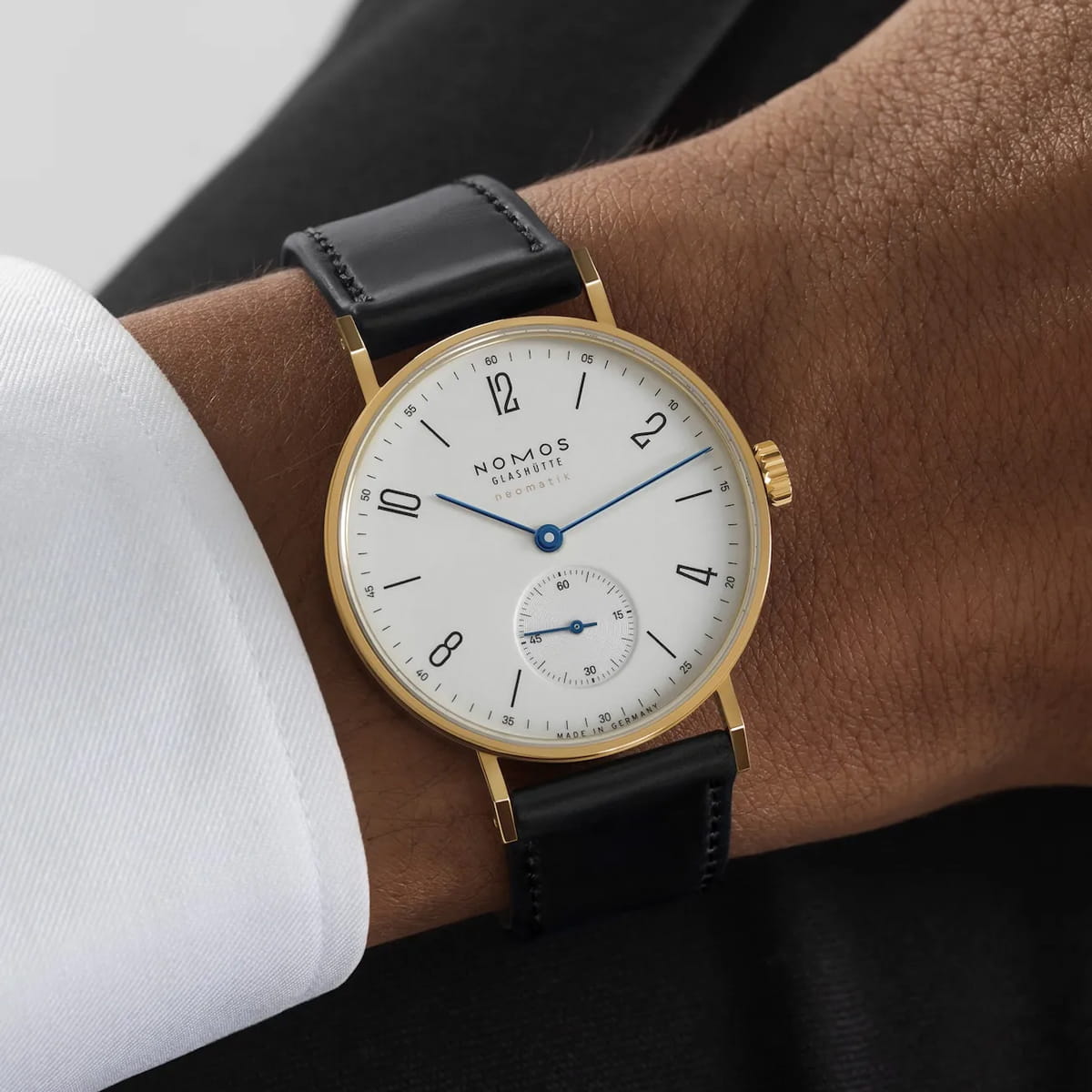 Eine NOMOS Tangente-Armbanduhr mit Goldgehäuse, weißem Zifferblatt, schwarzen Ziffern, blauen Zeigern und schwarzem Lederarmband, getragen am Handgelenk einer Person, die eine weiße Hemdmanschette trägt.