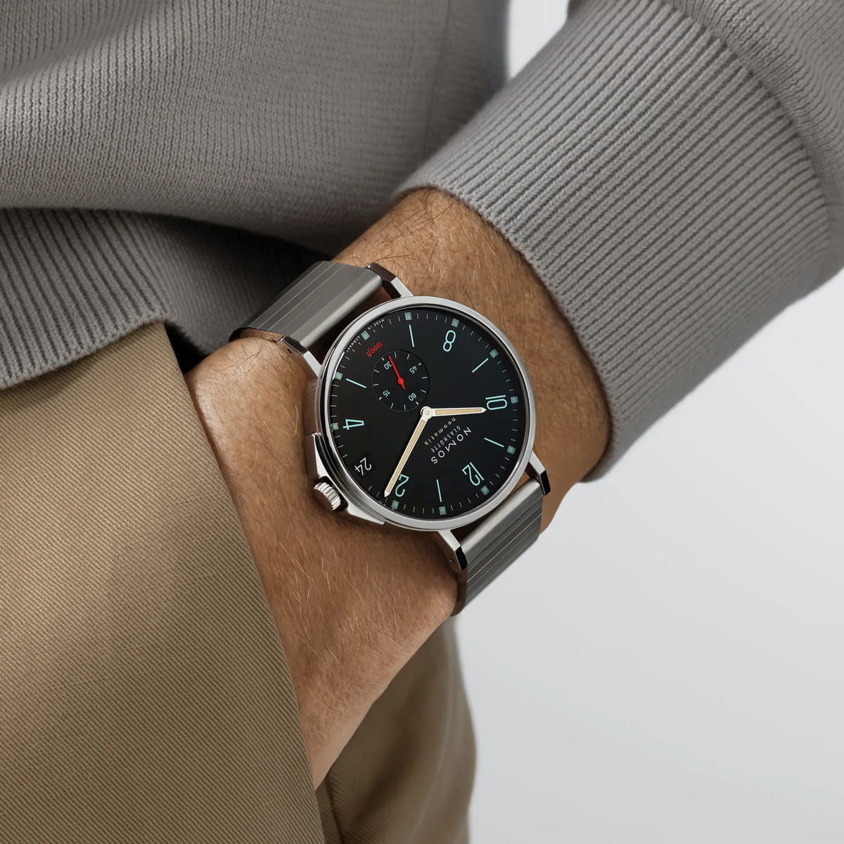 Eine Person mit einer silbernen NOMOS Tangente-Armbanduhr mit schwarzem Zifferblatt, blauen und weißen Ziffern und einem kleinen roten Ziffernblatt, bekleidet mit einem hellgrauen Pullover und einer hellbraunen Hose. Die Uhr hat ein minimalistisches, modernes Design.