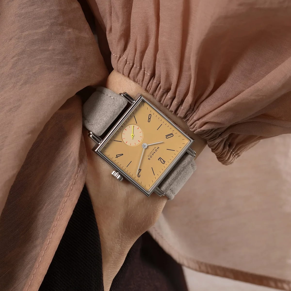 Eine Person trägt eine quadratische NOMOS-Tetra-Armbanduhr mit beigefarbenem Zifferblatt, zwei Hilfszifferblättern und einem hellgrauen Lederarmband. Die Uhr wird teilweise vom Ärmel einer weiten, hellbraunen Bluse verdeckt.