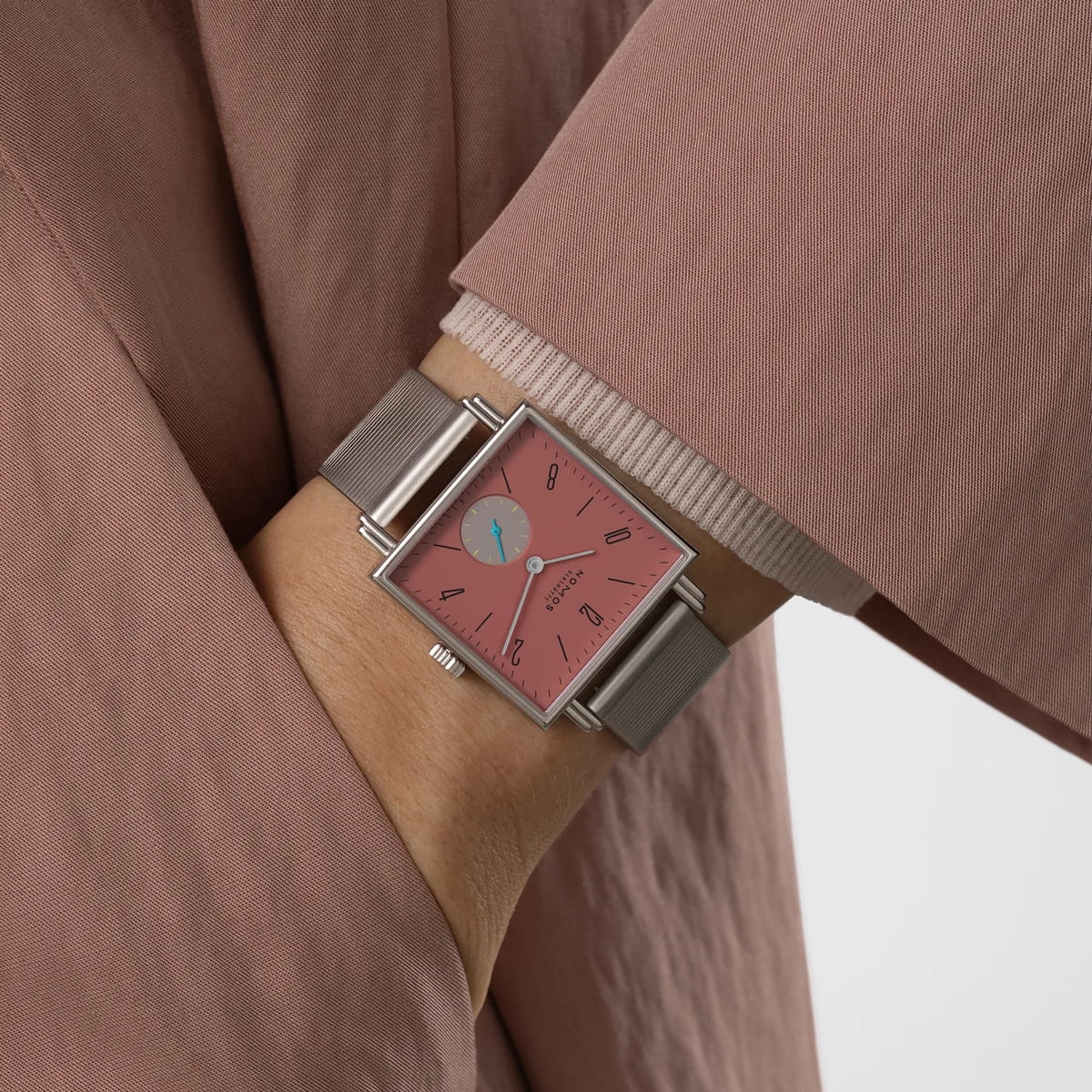 Eine Person, die eine silberne quadratische NOMOS-Tetra-Armbanduhr mit rosafarbenem Zifferblatt und blauem Sekundenzeiger trägt, bekleidet mit einem lila oder staubrosafarbenen Mantel, den Arm vor dem Körper verschränkt.