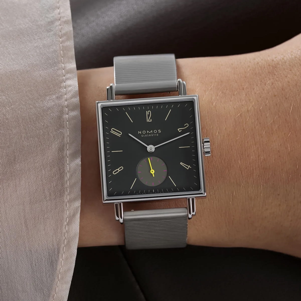 Eine quadratische NOMOS-Tetra-Armbanduhr mit schwarzem Zifferblatt, minimalistischen Indexen, kleiner Sekundenanzeige mit gelbem Zeiger und grauem Armband, die elegant am Handgelenk getragen wird.