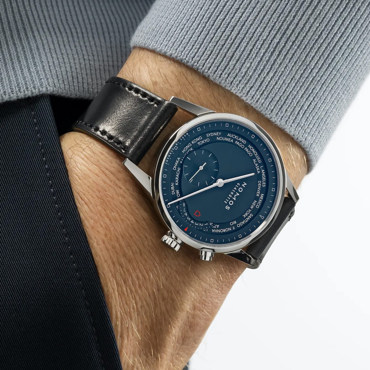 Eine Person trägt eine NOMOS-Zürich-Armbanduhr mit dunklem Lederband, blauem Zifferblatt, silberner Lünette und zwei Kronen, eine Hand in der Hosentasche und einen hellblauen Pullover sichtbar.