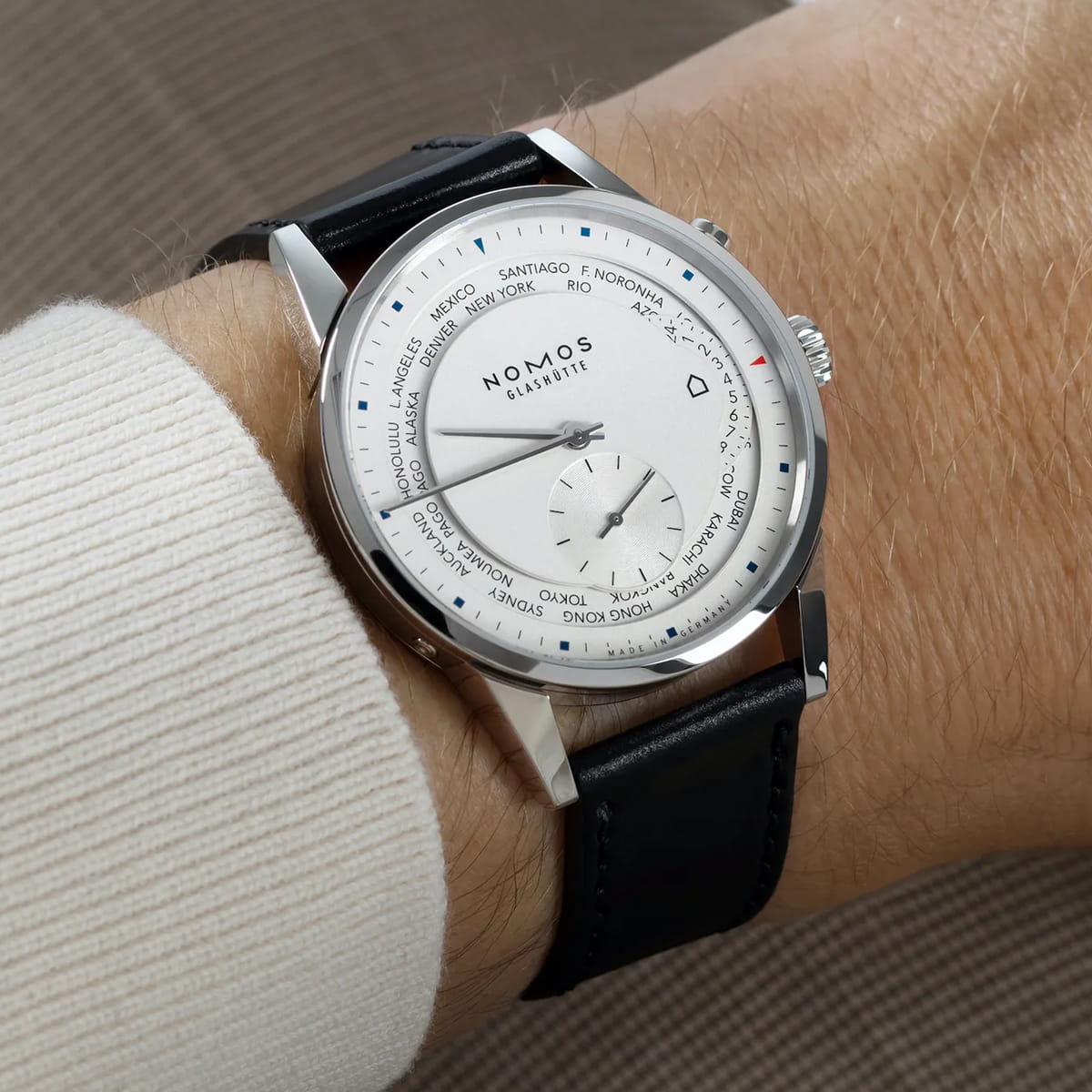 Nahaufnahme eines Handgelenks mit einer NOMOS Zürich Glashütte mit weißem Zifferblatt, schwarzem Lederband und Weltzeitstädten am äußeren Rand. Die Uhr besticht durch ihr modernes, elegantes Design.