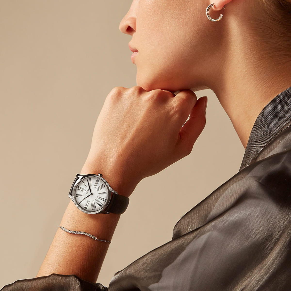 Eine Person, die eine silberne OMEGA DeVille-Armbanduhr mit weißem Zifferblatt und schwarzem Lederband, ein silbernes Armband und einen Ohrring trägt, bekleidet mit einem durchsichtigen schwarzen Oberteil, von der Seite gesehen vor einem beigen Hintergrund.