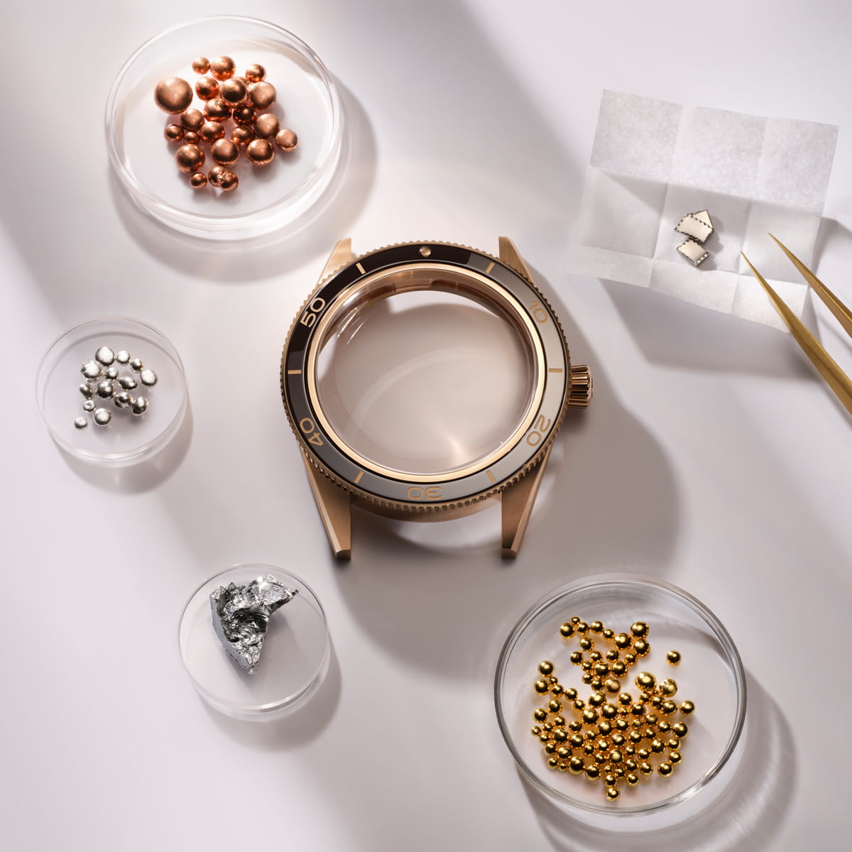 OMEGA © 2026 Ein goldenes OMEGA-Uhrengehäuse ist umgeben von kleinen Schalen mit Gold-, Silber- und Kupferperlen, einem Diamanten und zwei Metallfragmenten, daneben eine goldene Pinzette, die alle auf einer weißen Fläche angeordnet sind.