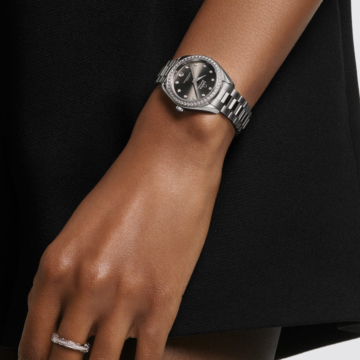 Eine Person mit mittlerem Hautton trägt eine silberne OMEGA Aqua Terra Uhr mit schwarzem Zifferblatt und Diamantenakzenten am linken Handgelenk, zusammen mit einem silbernen Ring, zu einem schlichten schwarzen Outfit.