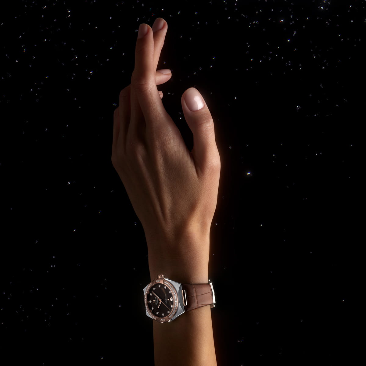 Eine Hand, die eine OMEGA Constellation Luxusarmbanduhr trägt, erhebt sich vor einem dunklen Hintergrund mit verstreuten Lichtpunkten, die an Sterne erinnern. Die Finger sind sanft geschwungen, und die Uhr hat ein dunkles Zifferblatt mit einem Metallarmband.