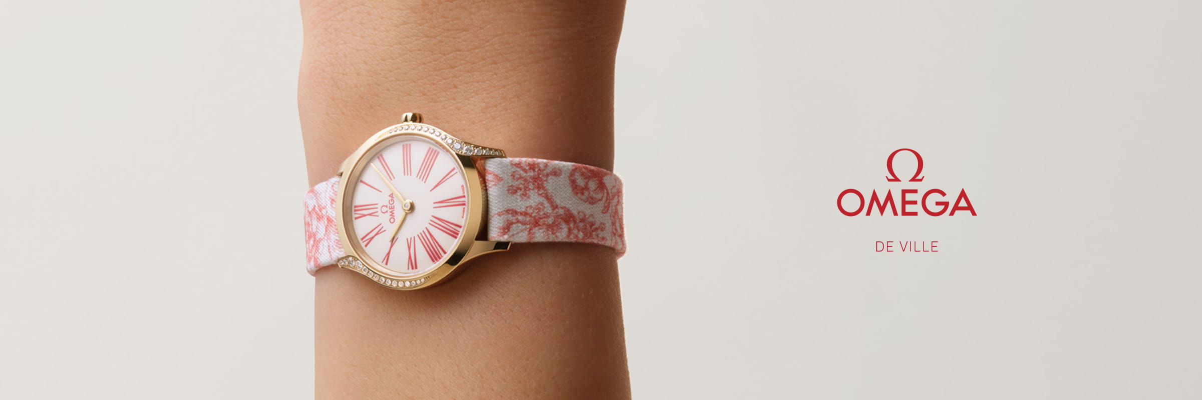 Eine goldene OMEGA DeVille Armbanduhr mit einem rosa und weißen, blumengemusterten Armband wird an einem Handgelenk getragen. Auf dem Ziffernblatt sind elegante römische Ziffern in Rosa und das OMEGA-Logo auf der rechten Seite zu sehen.