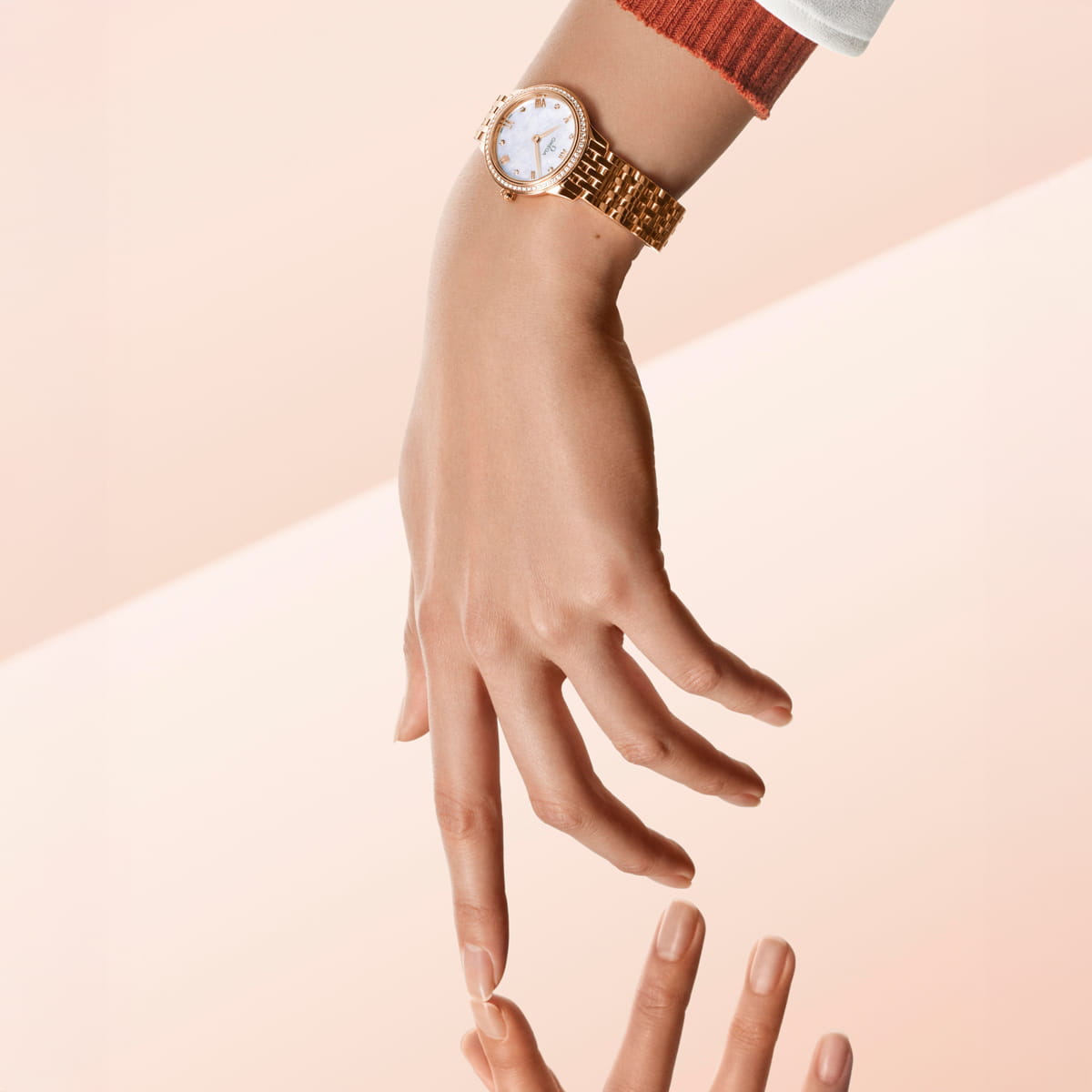 OMEGA © 2026 Eine Hand, die eine goldene OMEGA Prestige-Armbanduhr mit weißem Zifferblatt und Diamantenakzenten trägt, streckt die Finger aus und blickt auf ihr Spiegelbild auf einer glatten, neutralrosa Oberfläche.