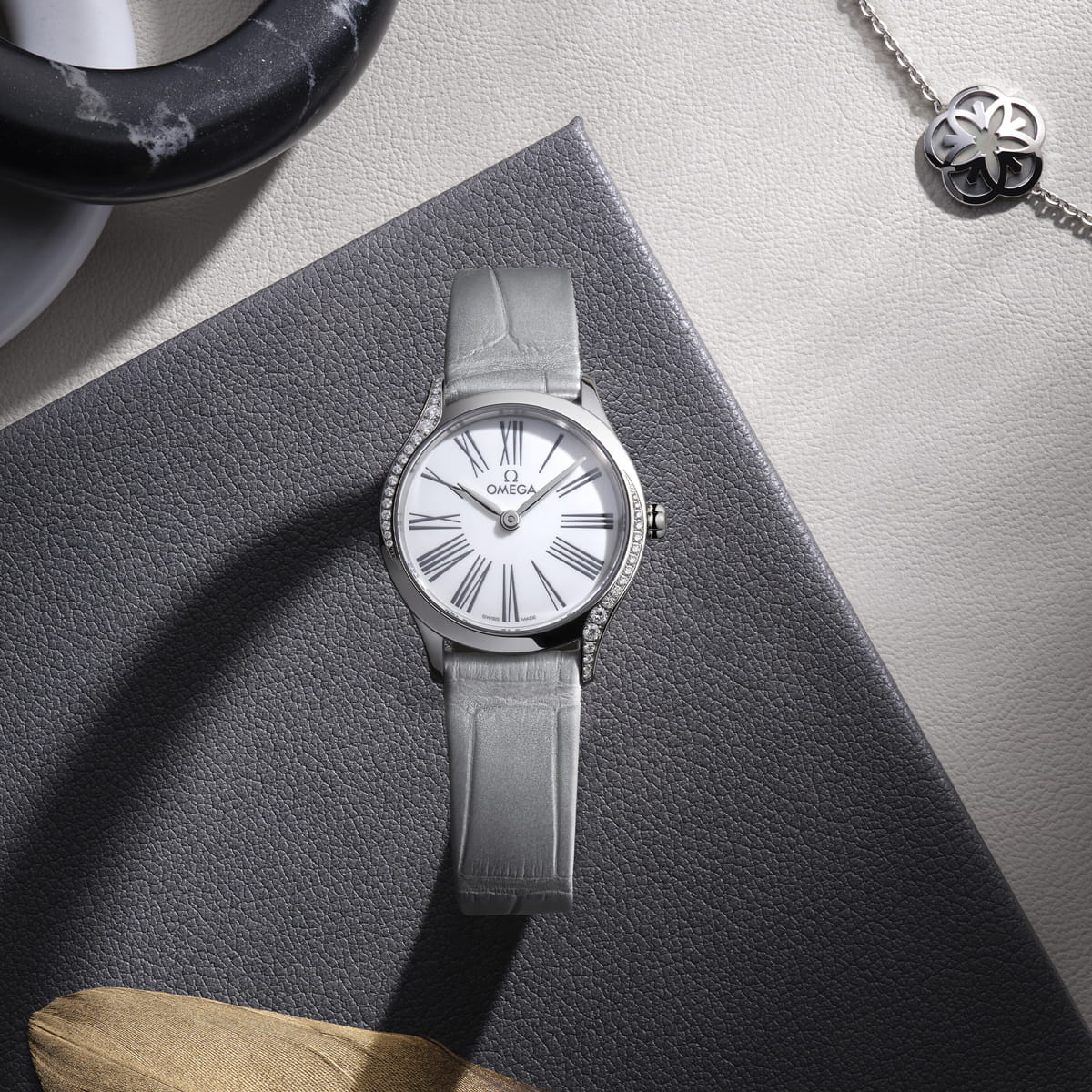 OMEGA © 2026 Eine silberne OMEGA Trésor-Armbanduhr mit weißem Ziffernblatt und römischen Ziffern ruht auf einer strukturierten grauen Oberfläche neben einer silbernen Halskette mit einem Kleeblattanhänger und einem Marmorobjekt.
