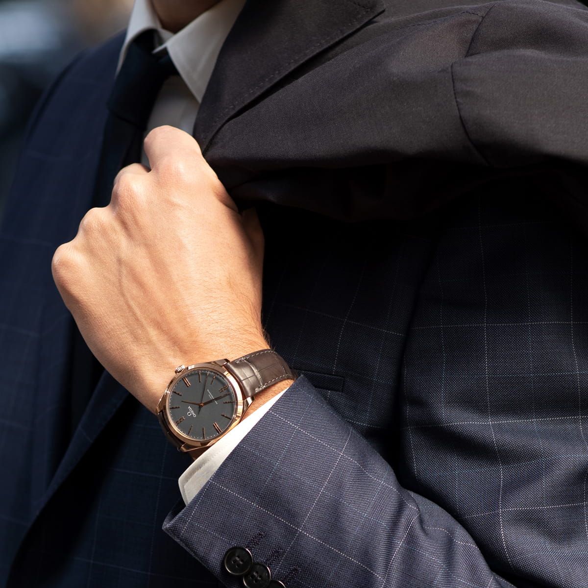 OMEGA © 2026 Eine Person in einem marineblau karierten Anzug mit Krawatte hält ihr Jackett über die Schulter und zeigt eine OMEGA Trésor Armbanduhr aus braunem Leder mit dunklem Zifferblatt an ihrem linken Handgelenk. Die Uhr und der Anzug sind auf dem Bild hervorgehoben.