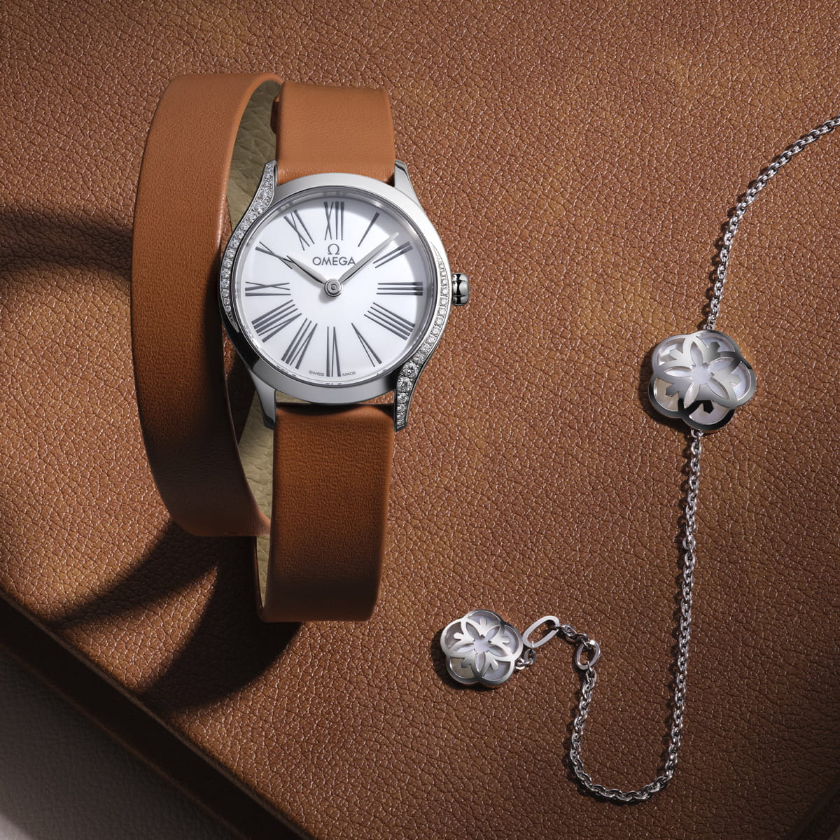 OMEGA © 2026 Eine silberne OMEGA Trésor-Armbanduhr mit weißem Zifferblatt und braunem Lederarmband liegt neben einem silbernen Kleeblattarmband auf einer strukturierten braunen Oberfläche.