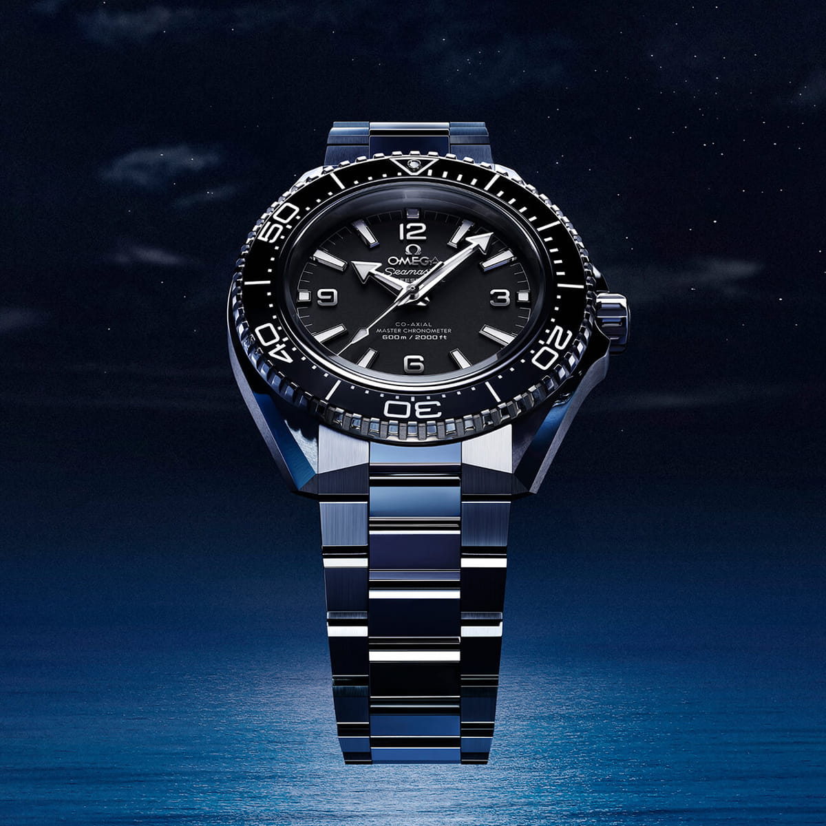 Eine luxuriöse OMEGA Planet Ocean Armbanduhr mit schwarzem Zifferblatt und silbernem Metallband wird vor einem dunklen, sternenklaren Nachthimmel und einem ruhigen blauen Ozean gezeigt. Die Uhr zeigt 10:08 an.