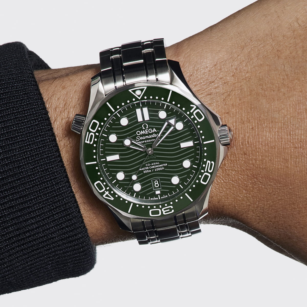 Nahaufnahme eines Handgelenks mit einer OMEGA Seamaster Uhr mit grünem Zifferblatt, fetten weißen Markierungen, silbernen Zeigern und einem Edelstahlarmband. Die Trägerin trägt ein schwarzes langärmeliges Oberteil.