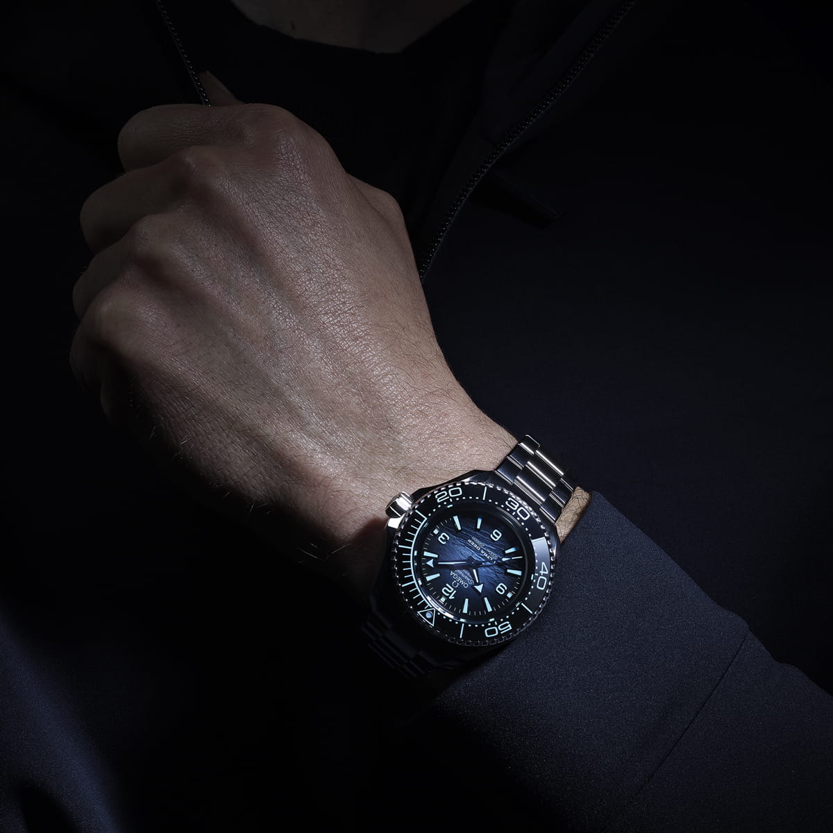 Eine Nahaufnahme der Hand und des Handgelenks einer Person, die eine OMEGA Seamaster trägt, eine analoge Uhr aus Edelstahl mit schwarzer Lünette und leuchtenden blauen Markierungen, vor einem dunklen Hintergrund.