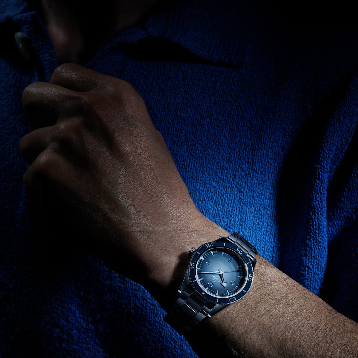 OMEGA © 2026 Eine Person rückt den Kragen eines blauen strukturierten Hemdes zurecht. Sie trägt eine OMEGA Seamaster Heritage mit blauem Zifferblatt, das unter fokussierter Beleuchtung vor einem dunklen Hintergrund dramatisch hervorgehoben wird.