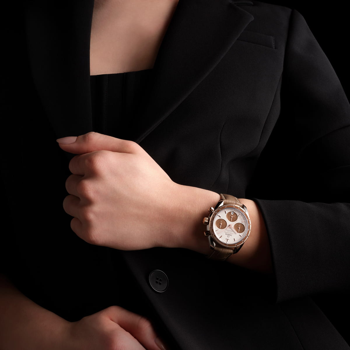 Eine Person in einem schwarzen Anzug hält sich mit einer Hand am Revers fest und trägt am anderen Handgelenk eine OMEGA Speedmaster 38 Goldarmbanduhr mit braunem Armband vor einem dunklen Hintergrund.