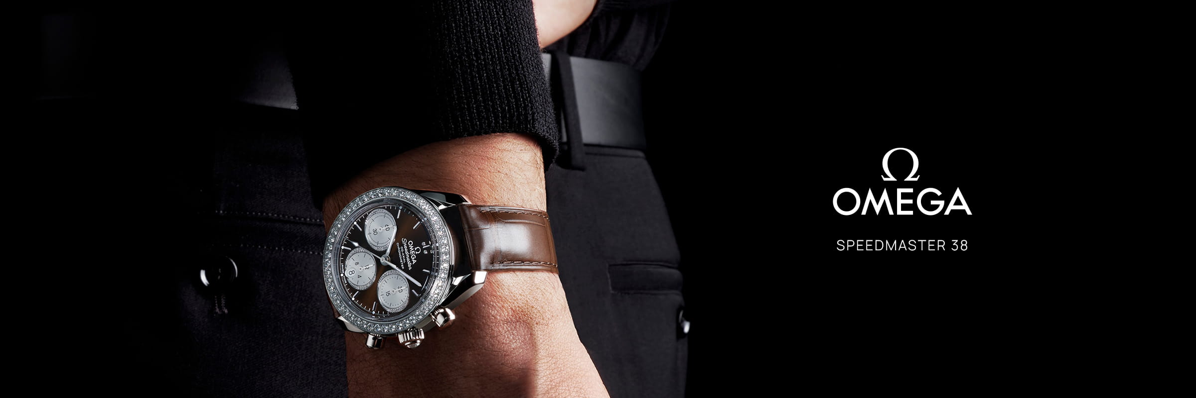 Nahaufnahme eines Handgelenks mit der OMEGA Speedmaster 38 Uhr an einem braunen Lederarmband. Der dunkle Hintergrund hebt das Omega-Logo und den Schriftzug "Speedmaster 38" auf der rechten Seite hervor.