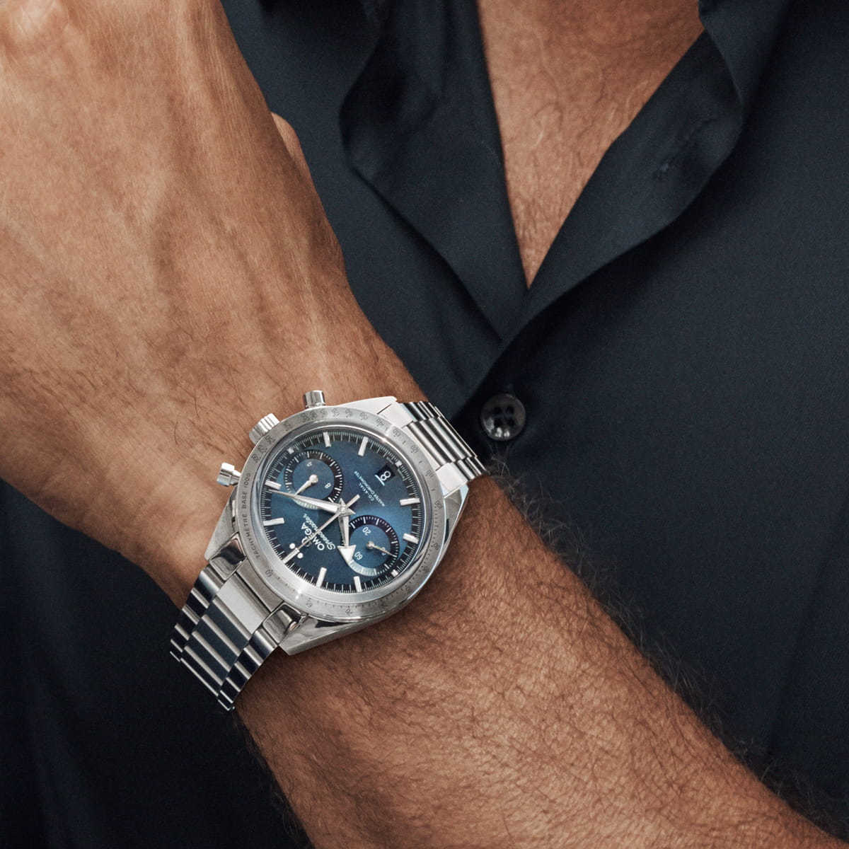 OMEGA © 2026 Eine Nahaufnahme des Handgelenks einer Person, die eine silberne Omega Speedmaster Chronographenuhr mit blauem Zifferblatt, mehreren Ziffernblättern und einem Metallgliederband trägt. Die Person ist mit einem kurzärmeligen, dunklen Hemd mit Knöpfen bekleidet.