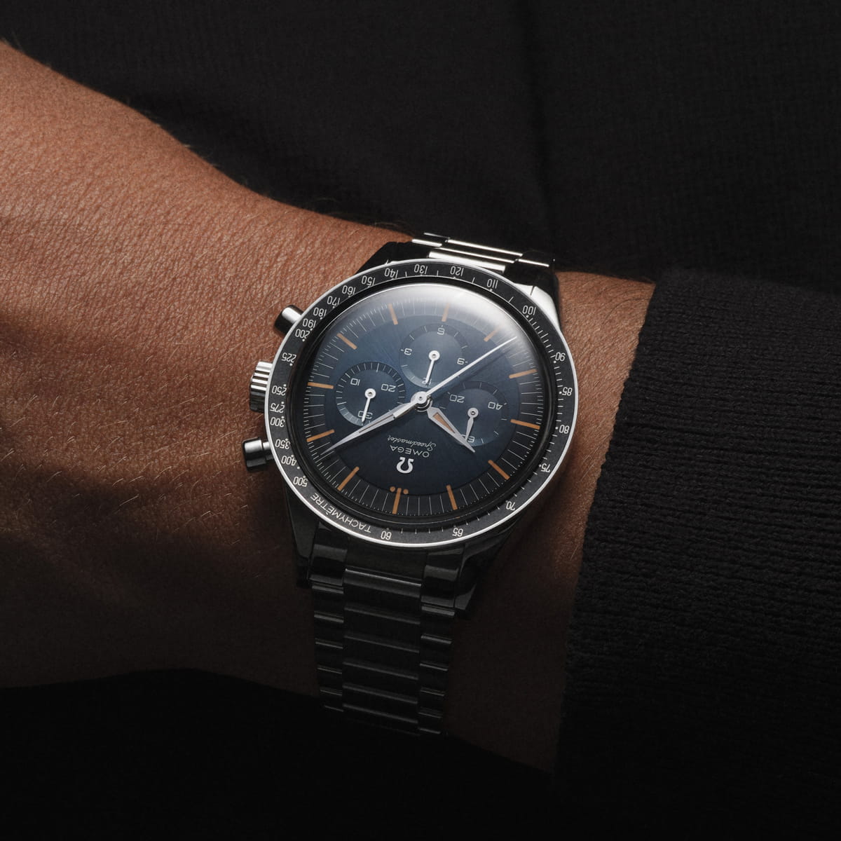 OMEGA © 2026 Nahaufnahme eines Handgelenks mit einer OMEGA Speedmaster Heritage Chronographenuhr aus Edelstahl mit schwarzem Zifferblatt, orangefarbenen Stundenmarkierungen und drei Hilfszifferblättern vor einem dunklen Hintergrund.