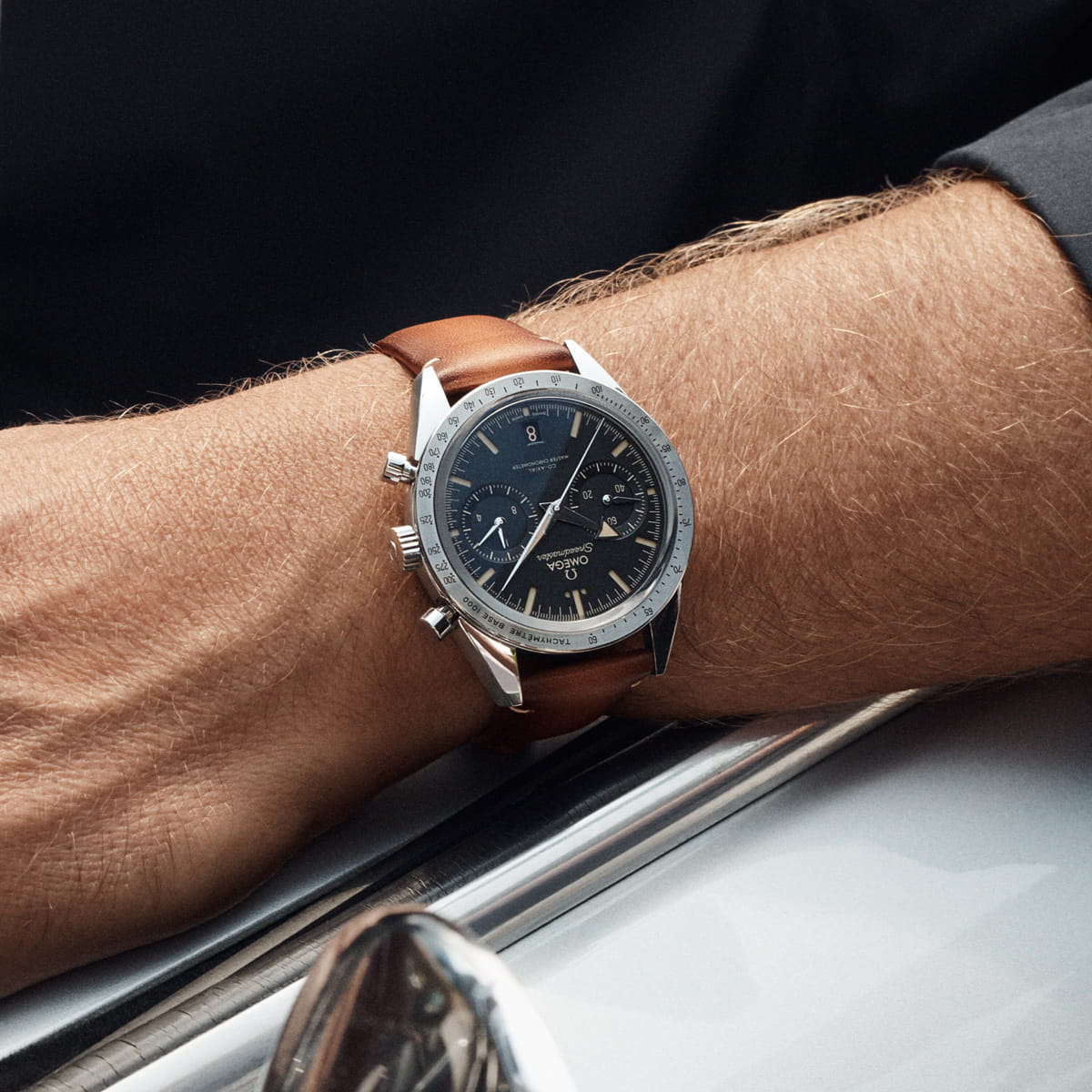 OMEGA © 2026 Nahaufnahme des Handgelenks einer Person, die eine OMEGA Speedmaster Heritage Uhr mit braunem Lederarmband und schwarzem Zifferblatt trägt, die an der Seite eines hellen Autos liegt.