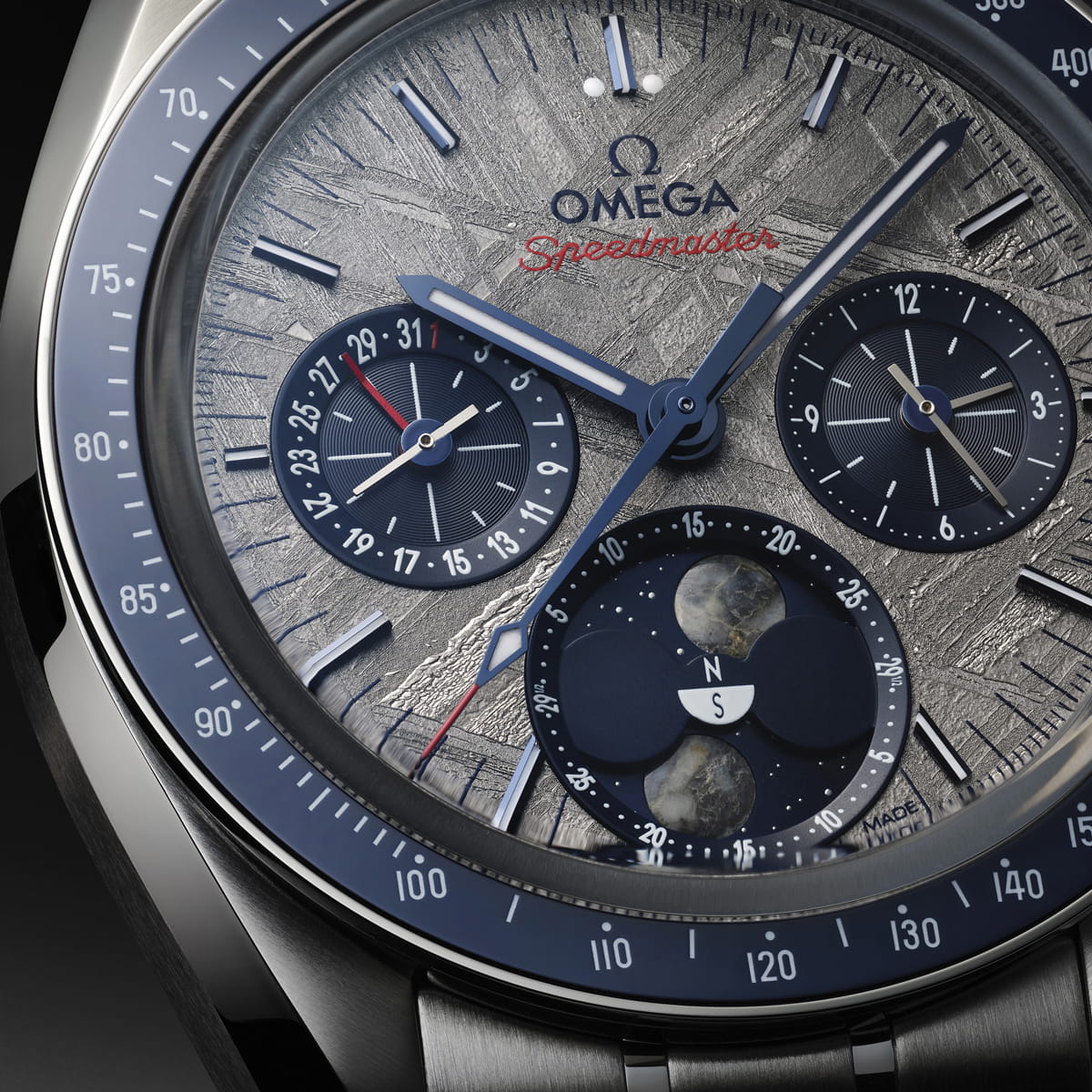 OMEGA © 2026 Nahaufnahme einer Omega Speedmaster-Armbanduhr im OMEGA Zwei Zähler-Design mit strukturiertem grauem Zifferblatt, blauen Hilfszifferblättern, Mondphasenanzeige und rotem Speedmaster"-Text. Die Uhr verfügt über aufwändige Details und ein poliertes Metallgehäuse.