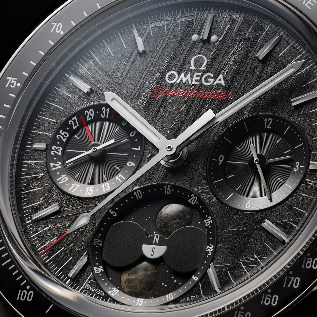 OMEGA © 2026 Nahaufnahme des Zifferblatts einer Omega Speedmaster OMEGA Zwei Zähler mit komplizierten Zifferblättern, strukturiertem dunklem Hintergrund und rotem "Speedmaster"-Schriftzug. Die Hilfszifferblätter zeigen das Datum, den Chronographen und eine Mondphasenanzeige mit einem Kompasssymbol.