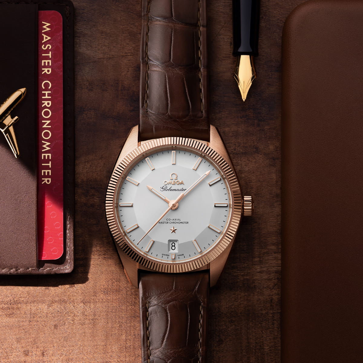 Eine OMEGA-Luxusarmbanduhr mit braunem Lederarmband und goldenem Gehäuse liegt auf einer hölzernen Unterlage, begleitet von einem Füllfederhalter, einem braunen Ledernotizbuch und einem roten Büchlein mit der Aufschrift "Master Chronometer".