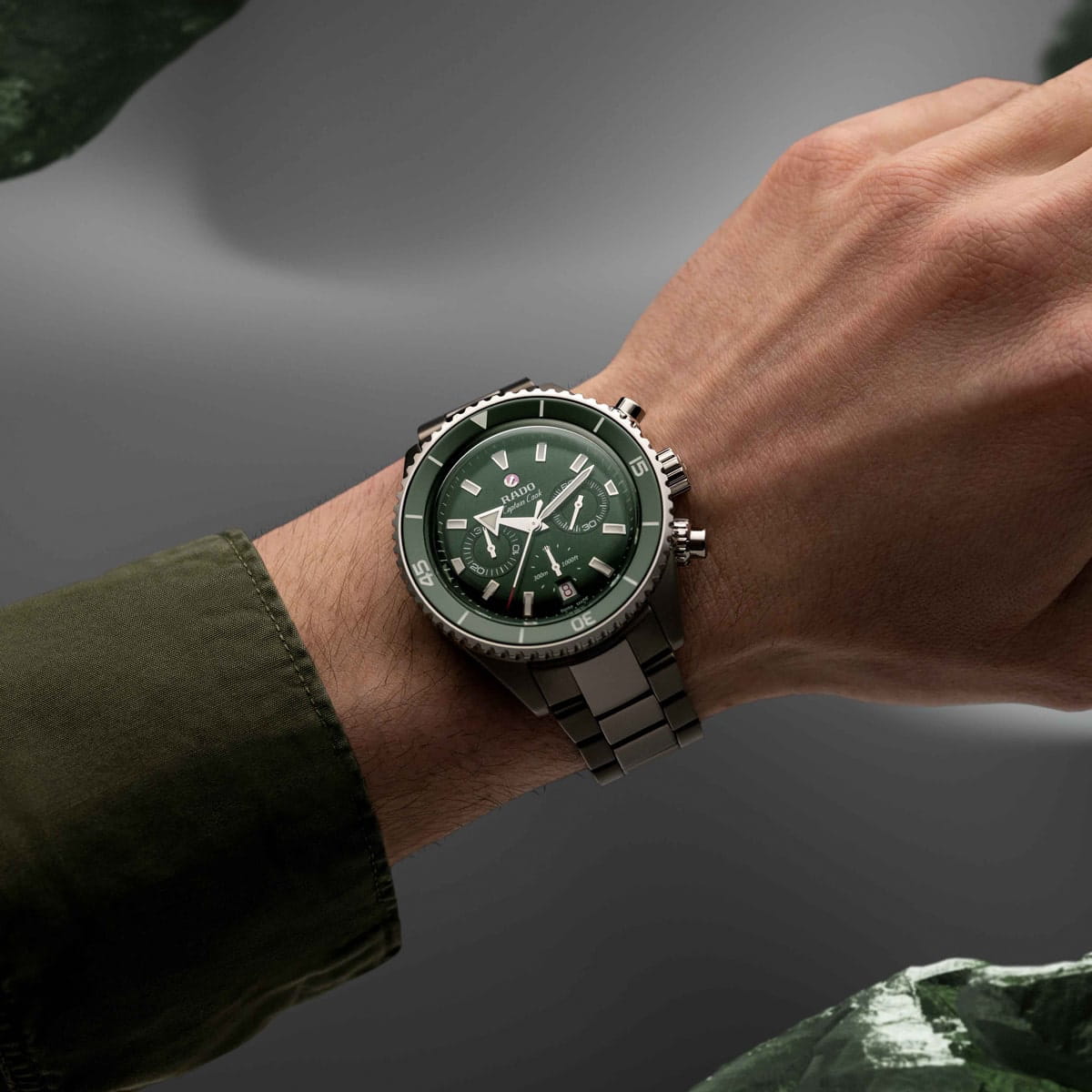 Eine Person trägt am Handgelenk eine silberne Rado Captain Cook Uhr mit grünem Zifferblatt und mehreren Ziffernblättern vor einem grauen Hintergrund. Die Person ist mit einem grünen langärmeligen Hemd bekleidet.