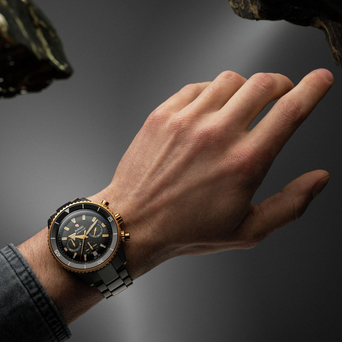Nahaufnahme der linken Hand einer Person, die eine luxuriöse zweifarbige Rado Captain Cook-Armbanduhr mit schwarz-goldenem Zifferblatt und silber-goldenem Band vor einem grauen Hintergrund mit dramatischer Beleuchtung trägt.