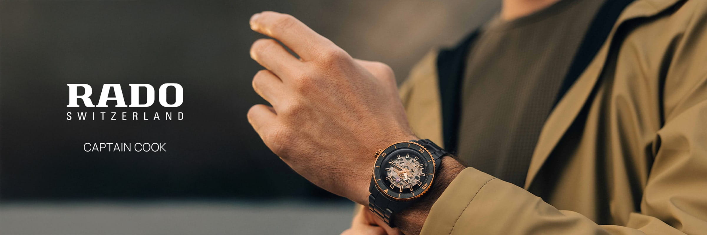 Eine Person, die eine hellbraune Jacke und ein grünes Hemd trägt, stellt ihre Rado Captain Cook-Armbanduhr ein. Das Bild zeigt den Text "RADO SWITZERLAND" und "CAPTAIN COOK" auf der linken Seite und hebt das klassische Rado Captain Cook Design hervor.