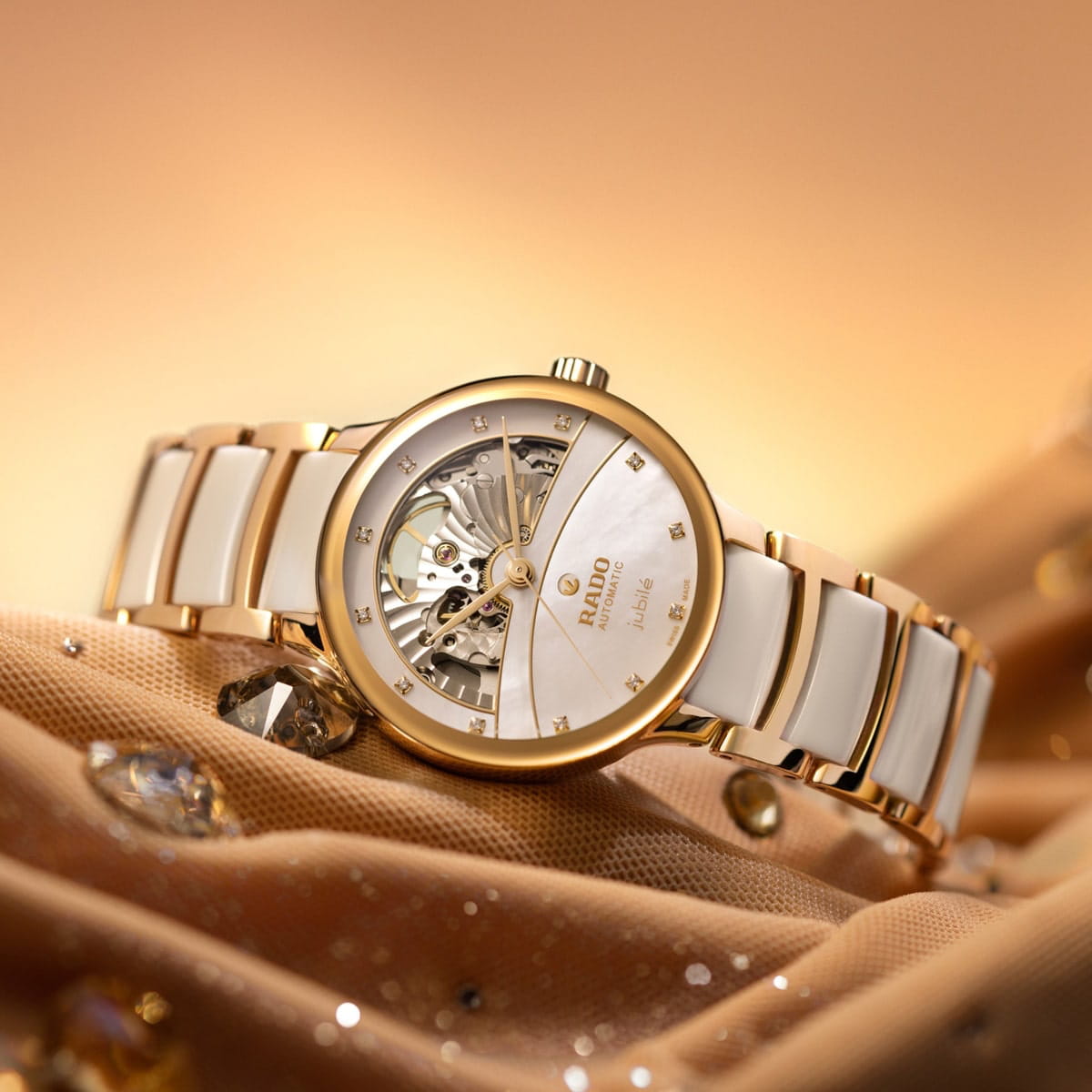 Eine goldene und weiße Rado Centrix Armbanduhr mit einem teilweise geöffneten Zifferblatt, das den Blick auf den inneren Mechanismus freigibt, präsentiert auf einem glitzernden, goldfarbenen Stoffhintergrund.