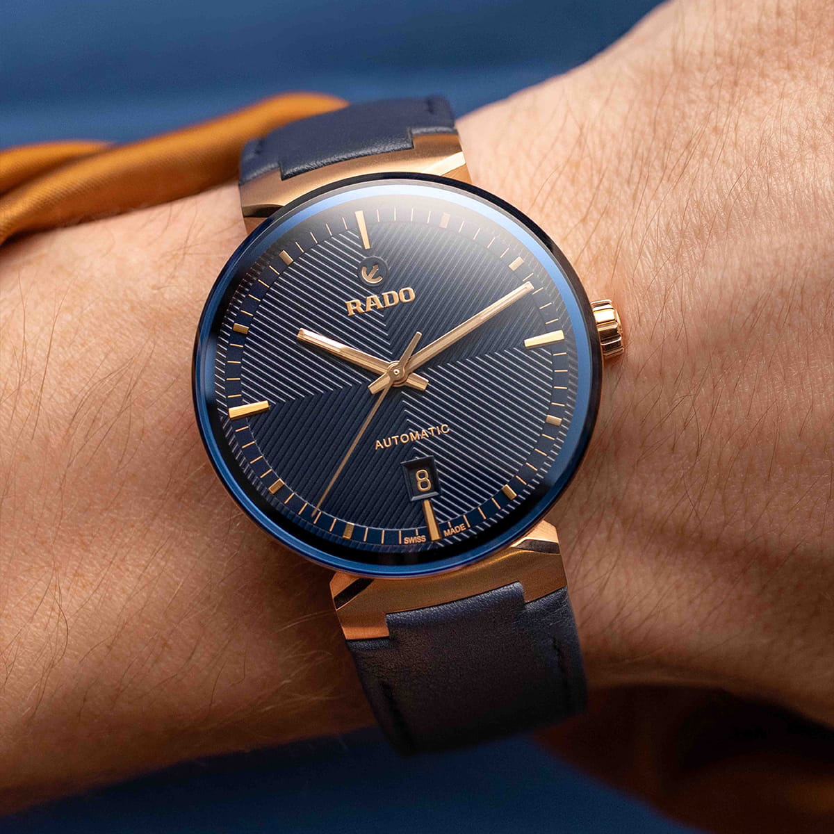 RADO © 2026 Eine Nahaufnahme einer Person, die eine Rado Florence Armbanduhr mit blauem Zifferblatt, goldenen Indexen und blauem Armband trägt. Die Uhr zeigt das Datum an und hat elegante goldene Akzente auf dem Gehäuse.