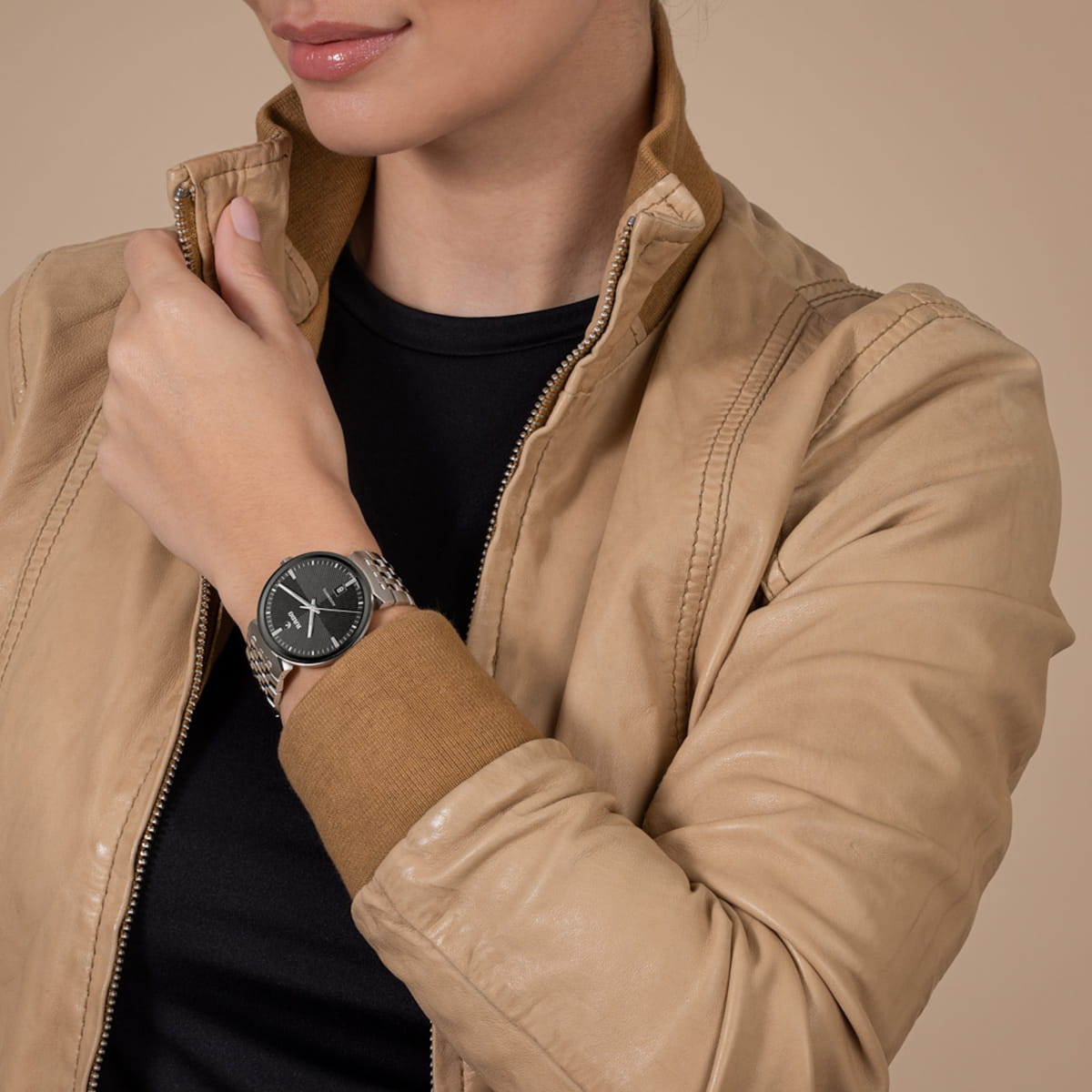 RADO © 2026 Eine Frau, die eine hellbraune Lederjacke und ein schwarzes Hemd trägt, zeigt eine silberne Armbanduhr von Rado Florence mit schwarzem Zifferblatt an ihrem linken Handgelenk vor einem schlichten beigen Hintergrund.