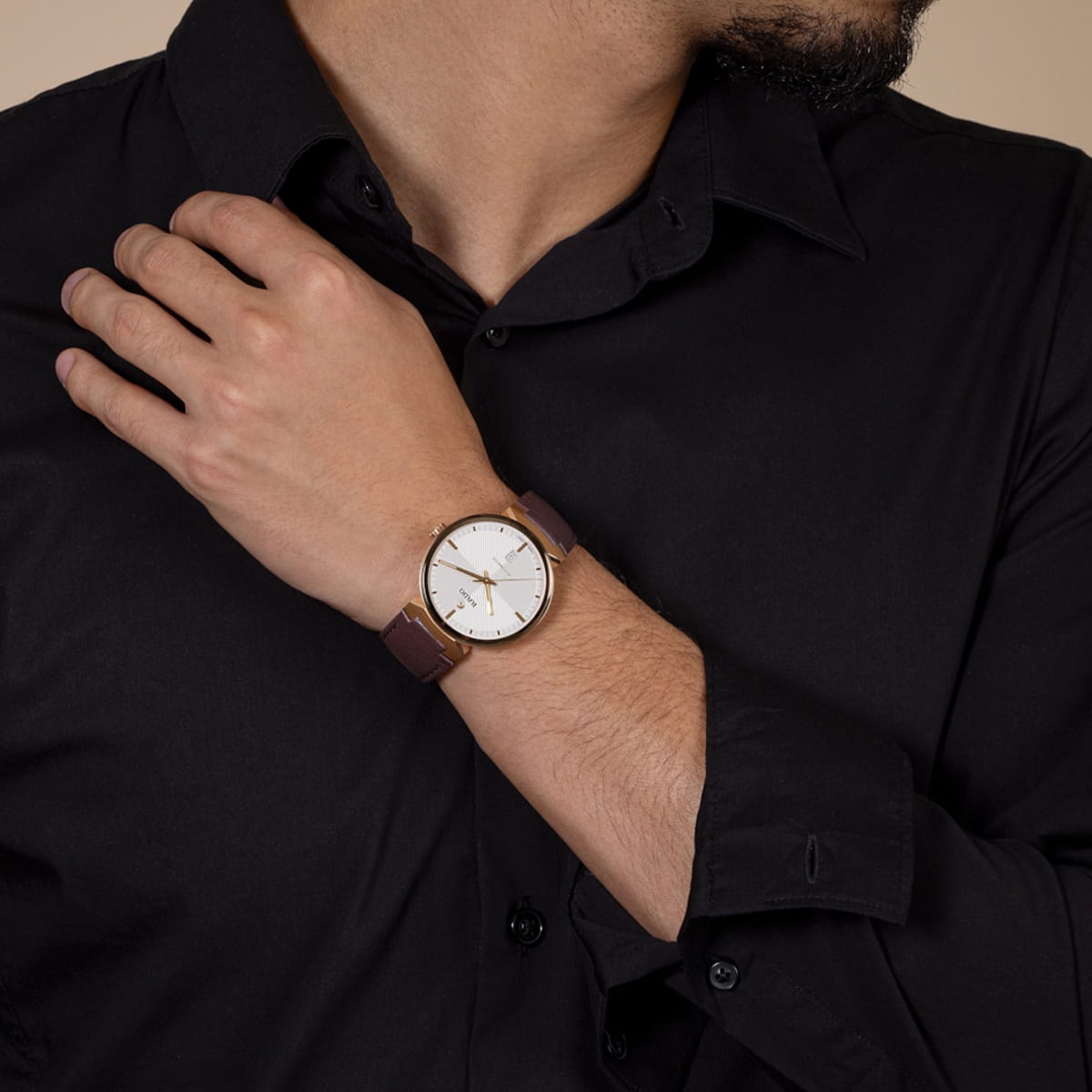 Eine Person, die ein schwarzes Button-up-Hemd trägt, zeigt eine Armbanduhr mit einem braunen Lederarmband und einem weißen, runden Zifferblatt und berührt mit einer Hand die gegenüberliegende Schulter.