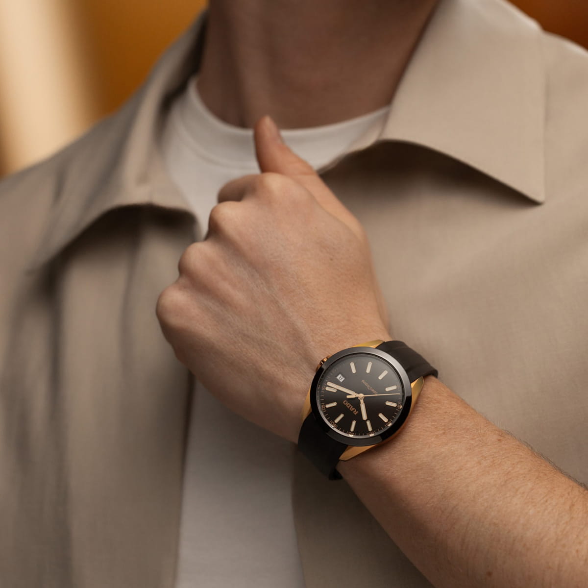 RADO © 2026 Eine Person, die ein beiges Hemd und ein weißes T-Shirt trägt, zeigt ihr Handgelenk, auf dem eine schwarz-goldene Rado Hyperchrome Analog-Armbanduhr mit einem einfachen, eleganten Design zu sehen ist. Das Ziffernblatt zeigt die Zeit 10:11 an.