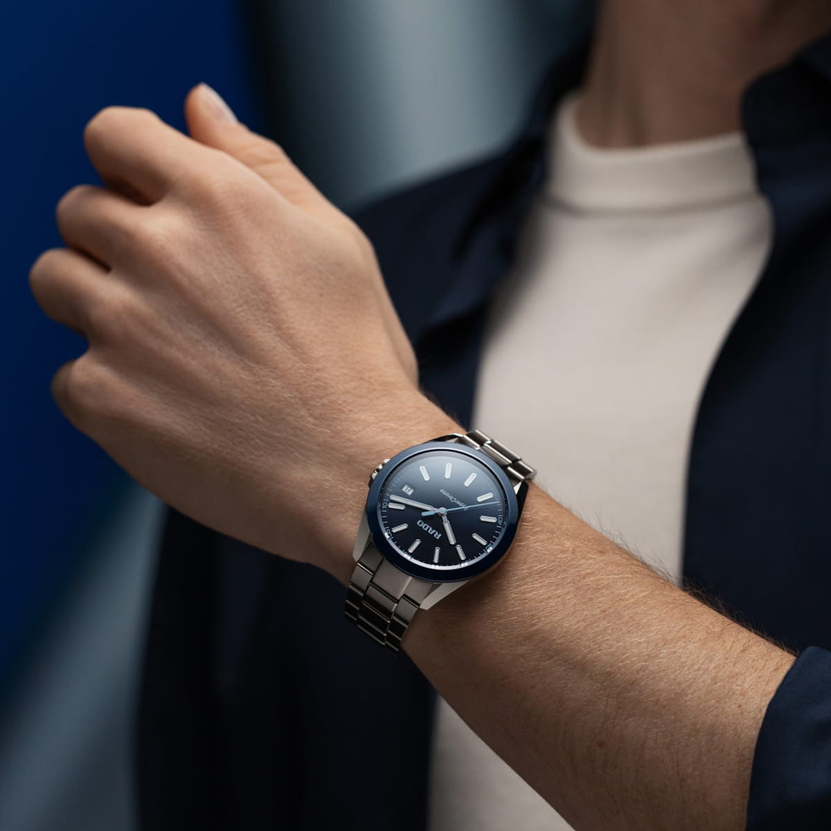 RADO © 2026 Eine Person trägt eine Rado Hyperchrome-Edelstahlarmbanduhr mit blauem Zifferblatt und silbernen Stundenmarkierungen am linken Handgelenk. Sie ist mit einem weißen Hemd und einer dunklen Jacke bekleidet, der Hintergrund ist leicht verschwommen.