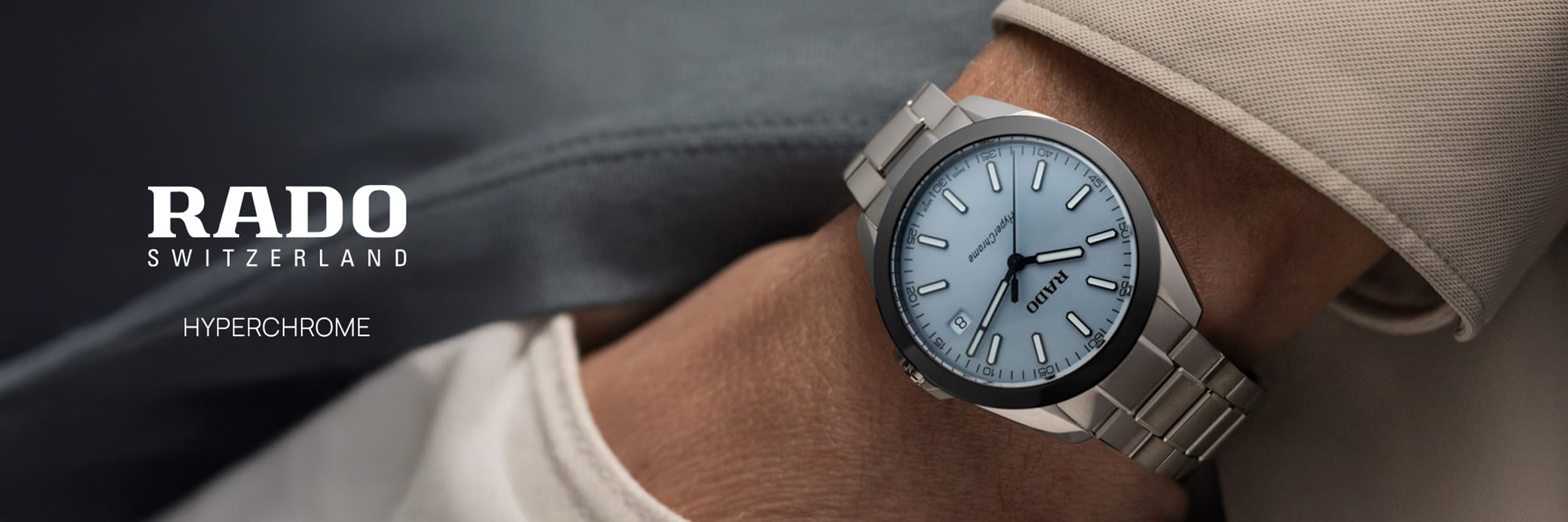 RADO © 2026 Eine Nahaufnahme einer Person, die eine elegante Rado Hyperchrome-Armbanduhr mit hellblauem Zifferblatt trägt, die die Zeit 10:10 anzeigt. Die Rado Hyperchrome hebt sich elegant von der neutralfarbenen Kleidung im Hintergrund ab.