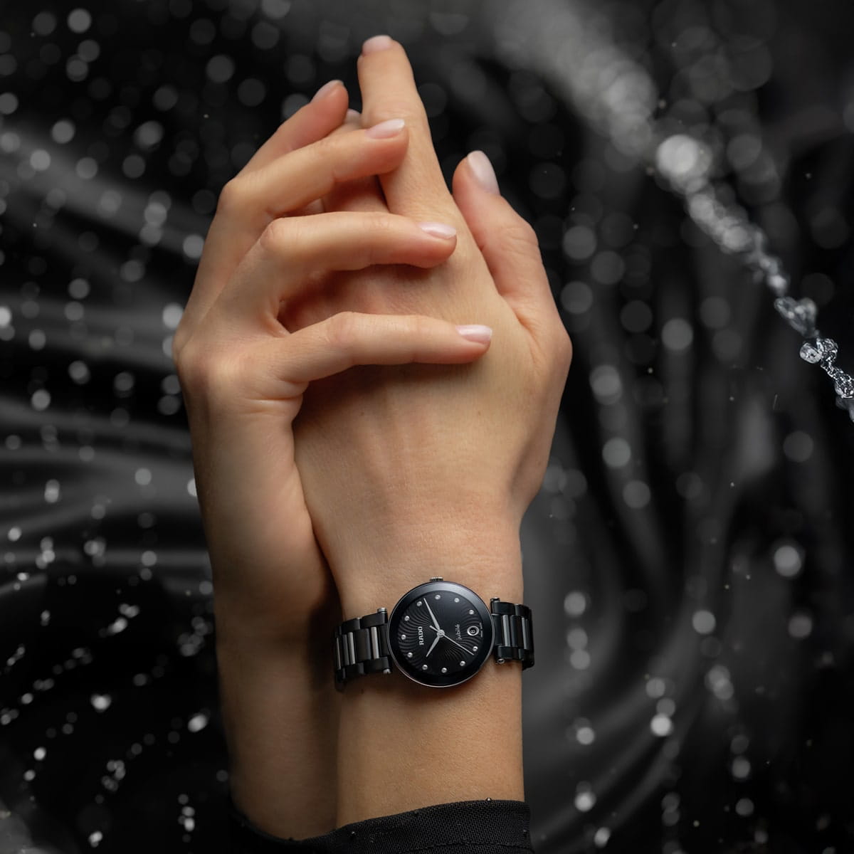 Eine Nahaufnahme von zwei ineinander verschränkten Händen, von denen eine eine elegante schwarze Rado LaCoupole Armbanduhr trägt, wobei Wassertropfen und ein dunkler, gewellter Hintergrund einen dynamischen, eleganten Effekt erzeugen.