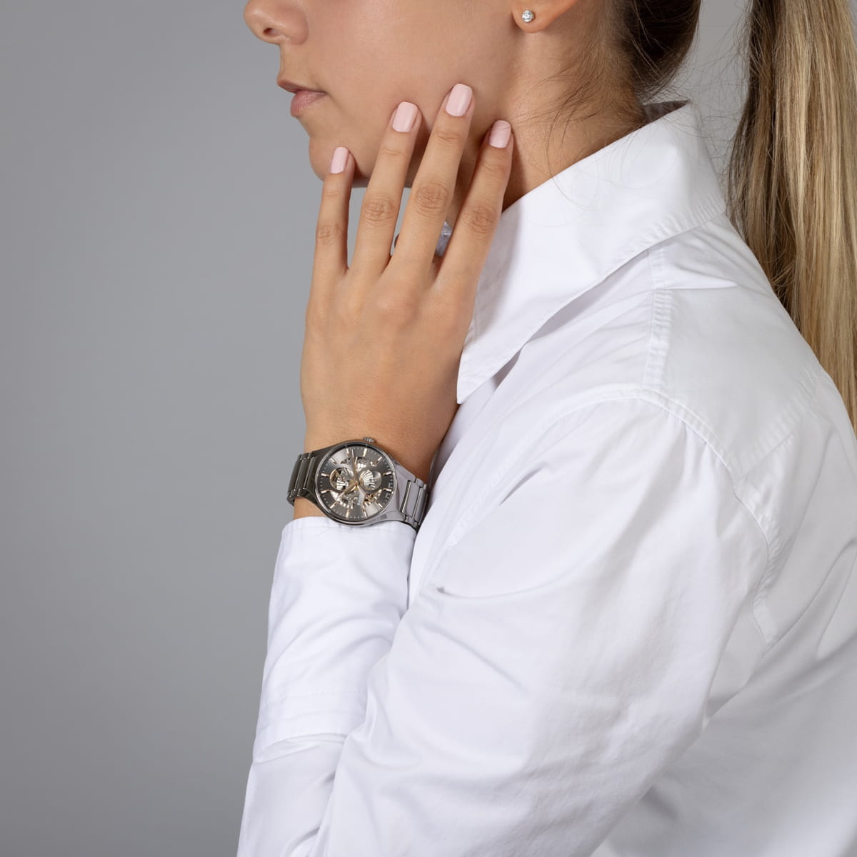 Eine Frau im weißen Hemd berührt mit der linken Hand ihr Gesicht und zeigt eine silberne Armbanduhr mit einem sichtbaren, komplizierten Mechanismus. Ihr blondes Haar ist zurückgebunden, sie hat hellrosa Nägel und einen Ohrstecker.