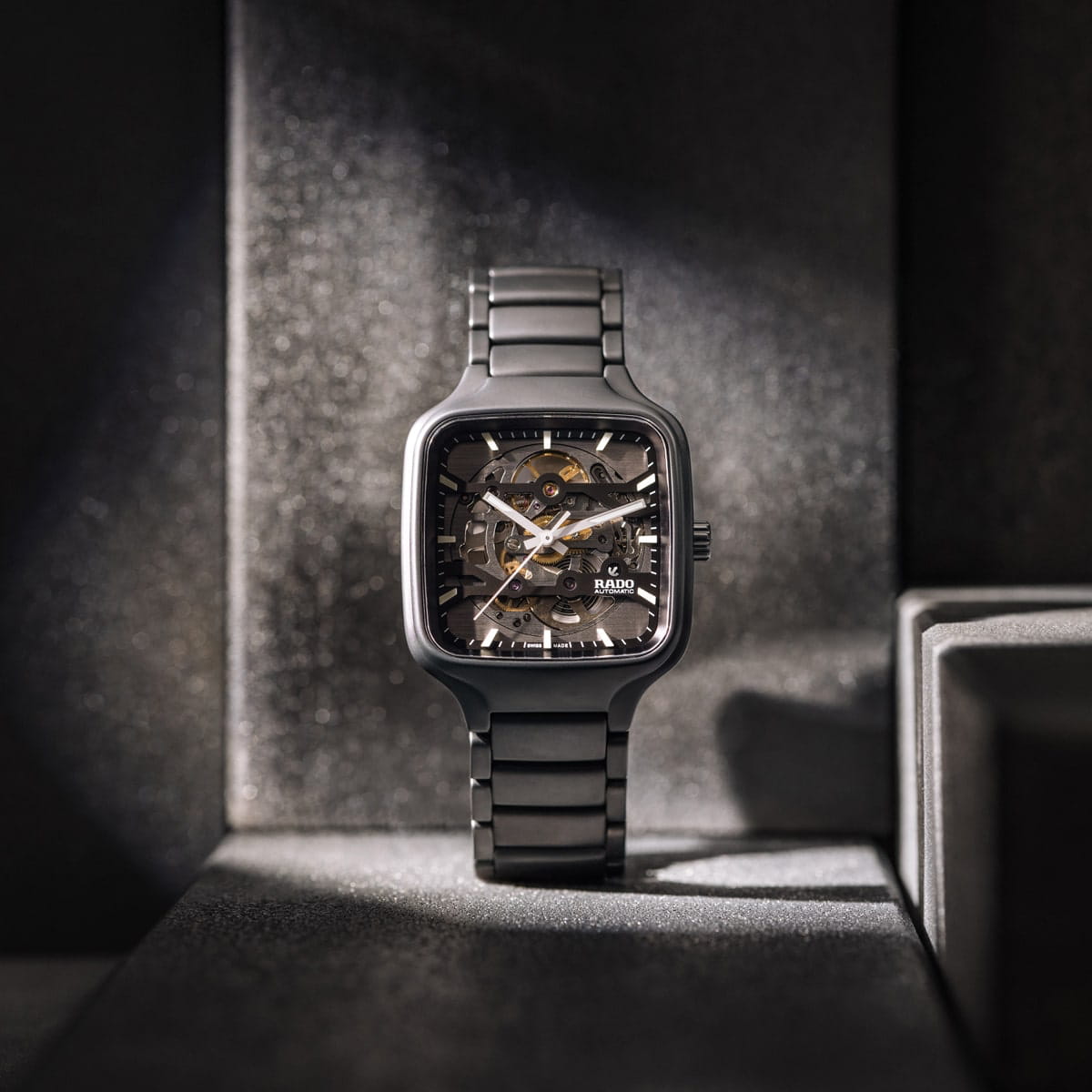 Eine Rado True Square Skelettuhr mit Metallarmband wird aufrecht in einer schwach beleuchteten, schattigen Umgebung präsentiert, um die komplizierten inneren Zahnräder und mechanischen Details hervorzuheben.
