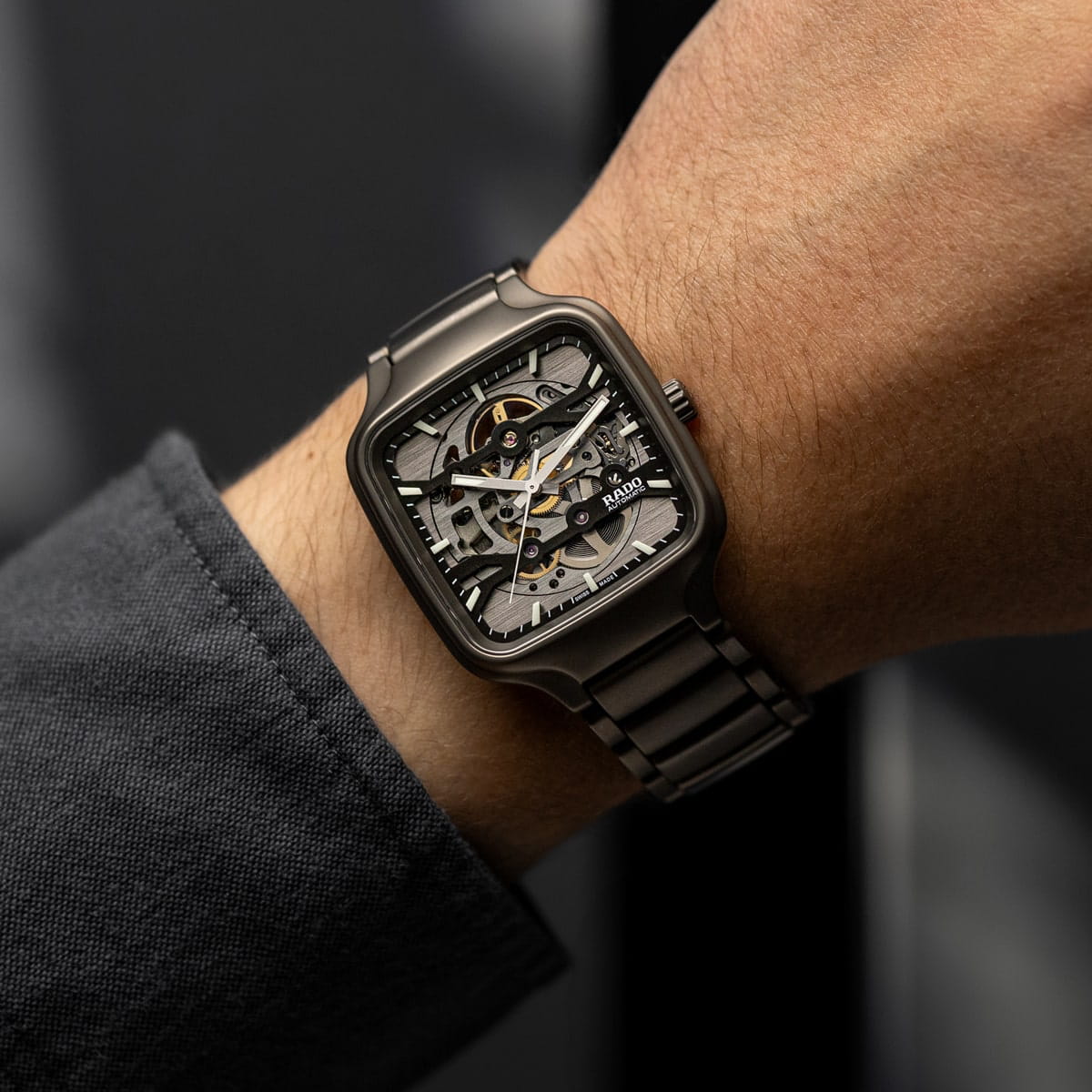 RADO © 2026 Nahaufnahme des Handgelenks einer Person, die eine quadratische, metallische Rado-Skelettuhr mit sichtbaren Zahnrädern und einem schwarzen Metallband trägt. Die Person ist mit einem dunklen langärmeligen Hemd bekleidet.