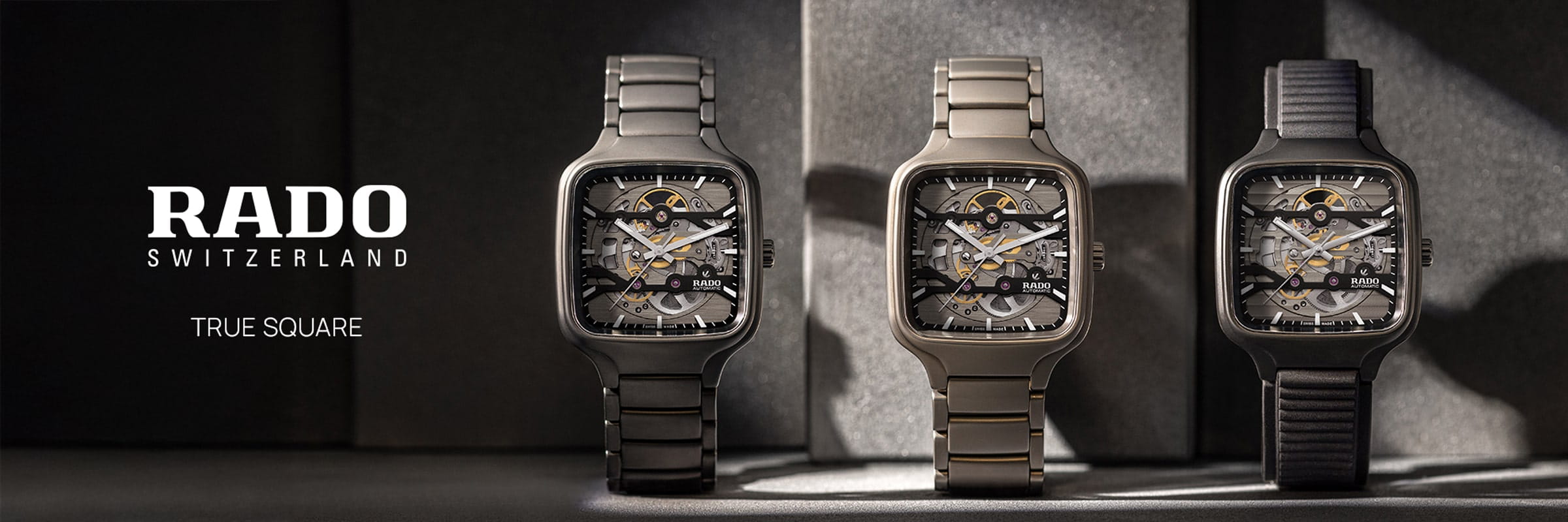 RADO © 2026 Drei Rado True Square Skelettuhren sind nebeneinander auf Ständern ausgestellt, jede mit einem quadratischen Gehäuse und sichtbarer Mechanik im Inneren, mit verschiedenen Metall- und Keramikbändern. Auf der linken Seite ist das Logo von Rado Switzerland zu sehen.