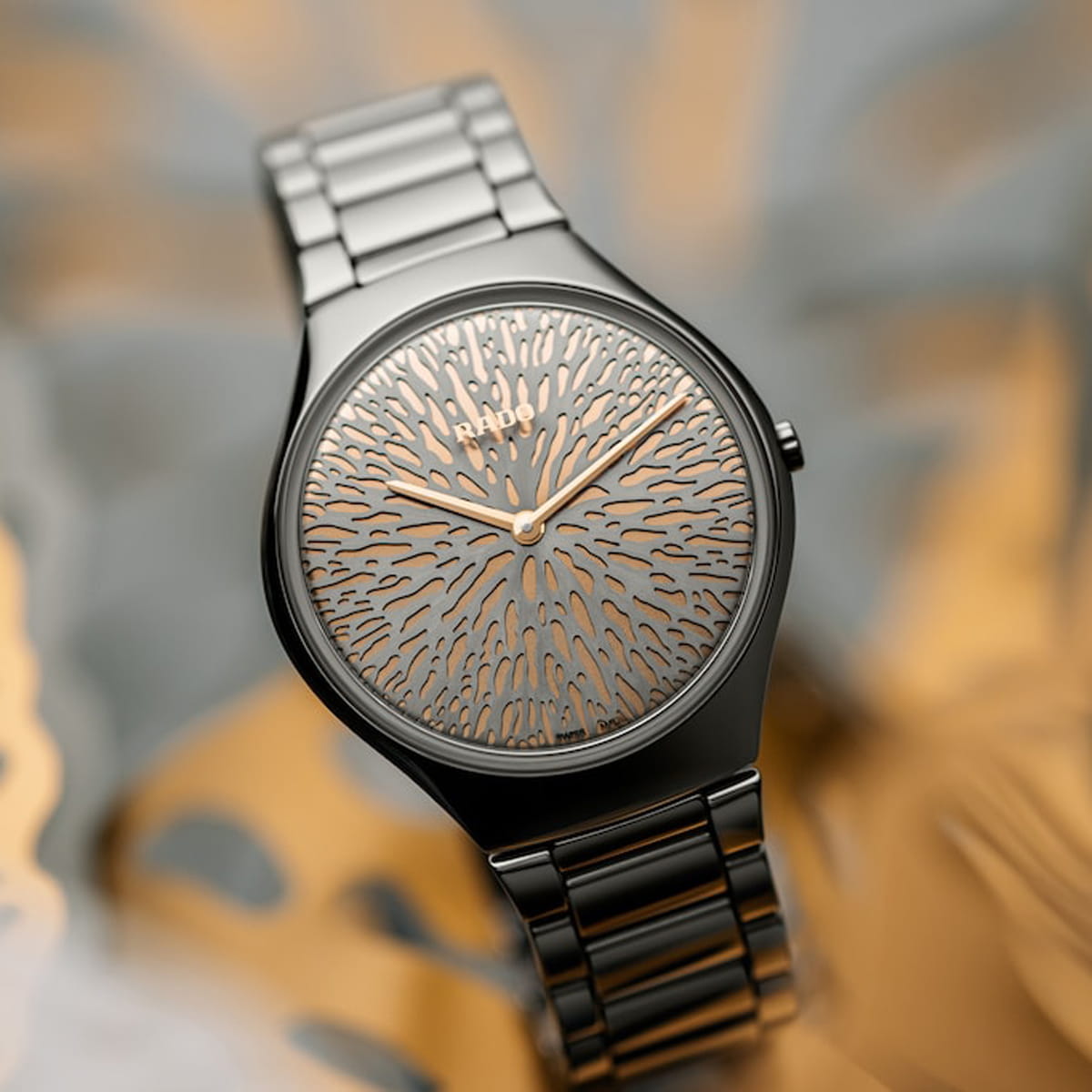 Eine elegante, metallische Armbanduhr mit einer gemusterten, korallenähnlichen Struktur auf dem Zifferblatt, die keine Ziffern aufweist. Die Zeiger und das Muster sind goldfarben, während das Armband und das Gehäuse aus glänzendem, dunklem Metall bestehen. Der Hintergrund ist abstrakt und unscharf.